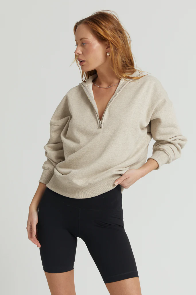 Woman wearing a beige zip-up sweatshirt and black shorts on a light gray background