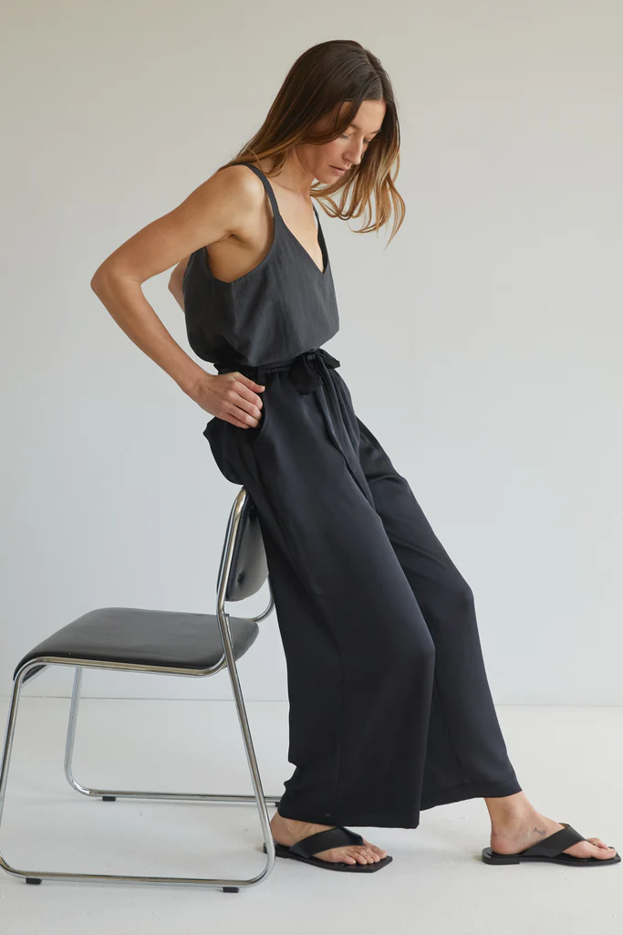 Woman in a black v neck top and black waist tie pants sitting on a stool against a white background