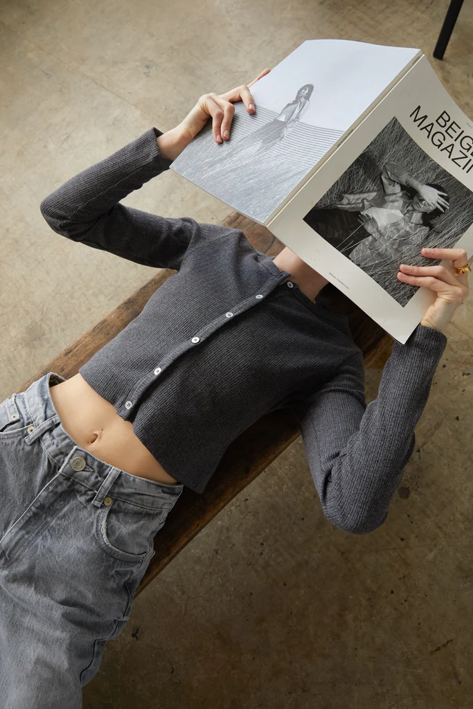 Woman wearing a gray sweater and jeans, holding a magazine over their face on a stone floor.