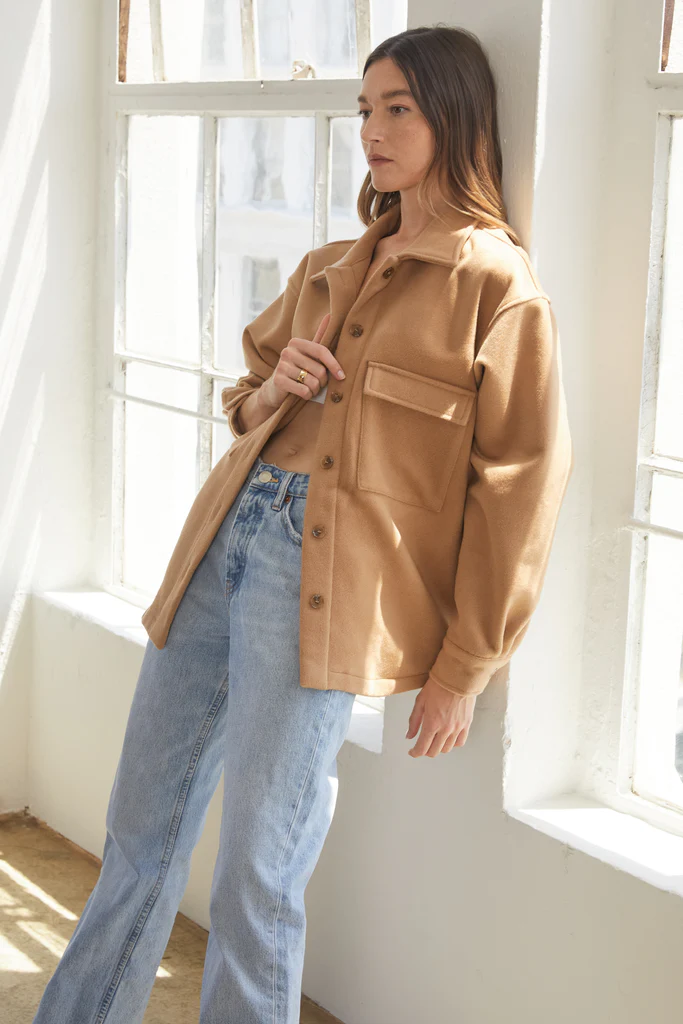 Woman wearing a tan jacket and blue jeans standing in a bright room with large windows.