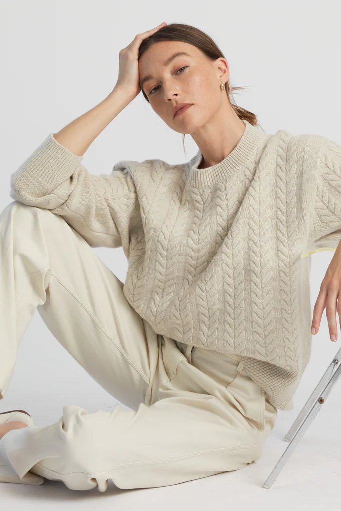 Woman wearing a beige cable knit sweater and pants sitting on a white surface.