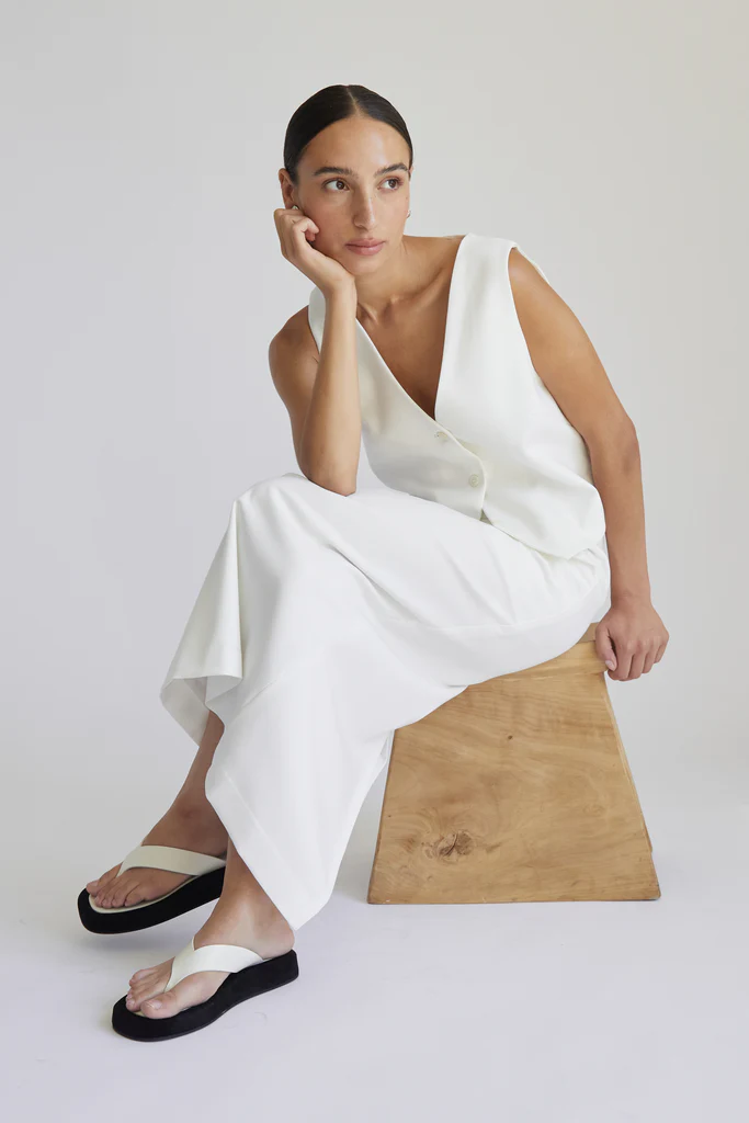 Woman in a white outfit sitting on a wooden stool against a plain background
