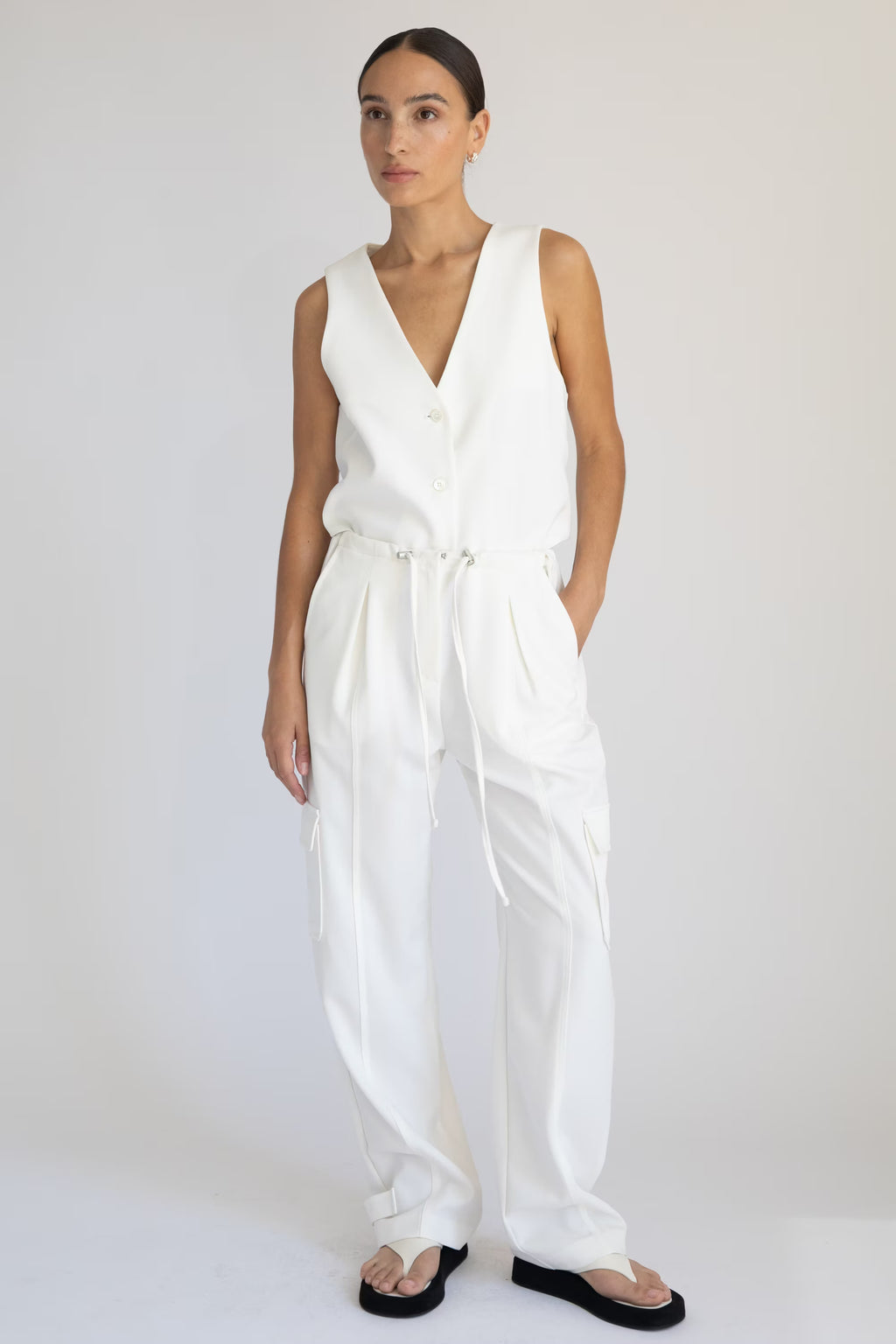 Woman wearing a white sleeveless vest and white cargo pants on a plain background