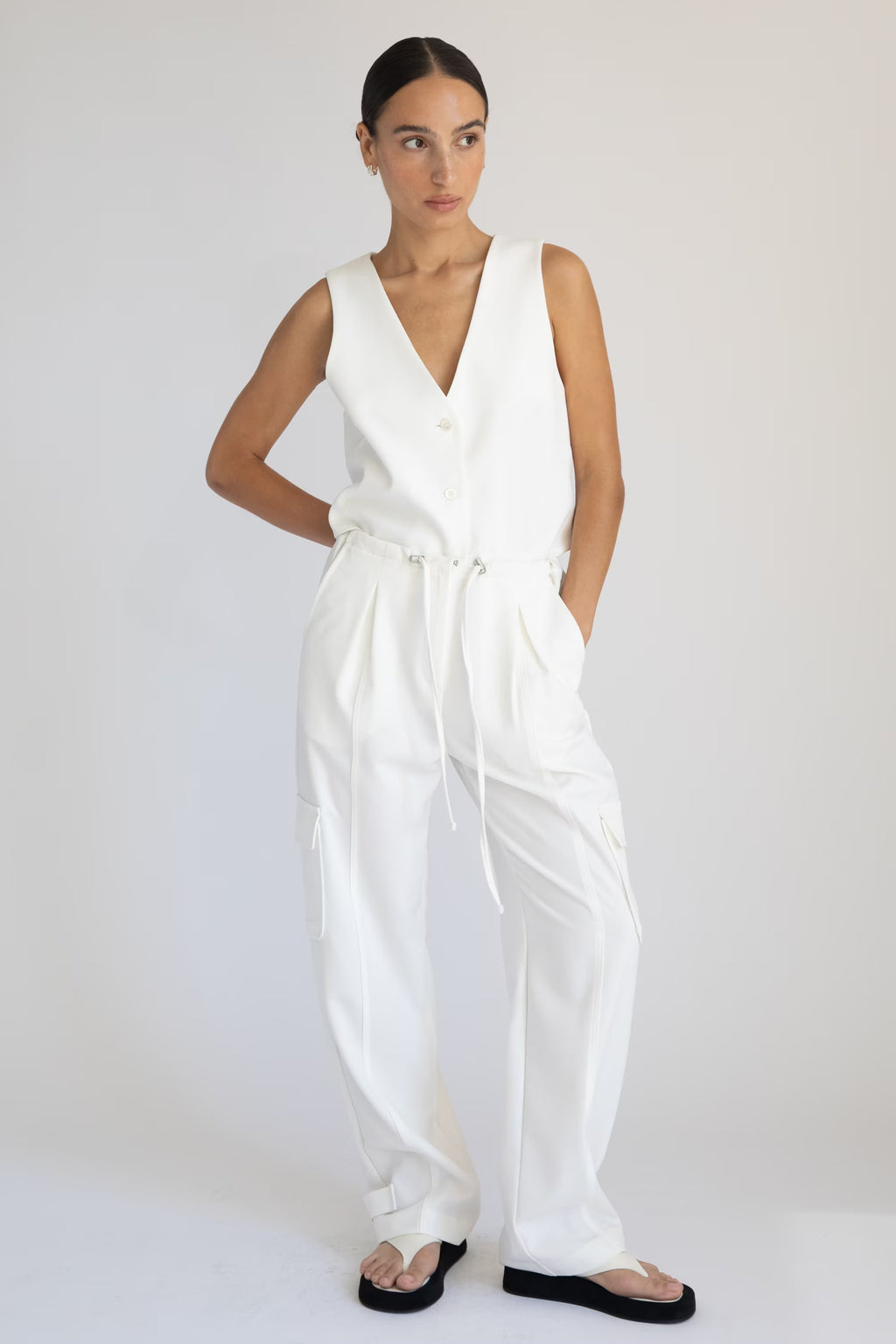 Woman wearing a white sleeveless vest and white cargo pants on a plain background