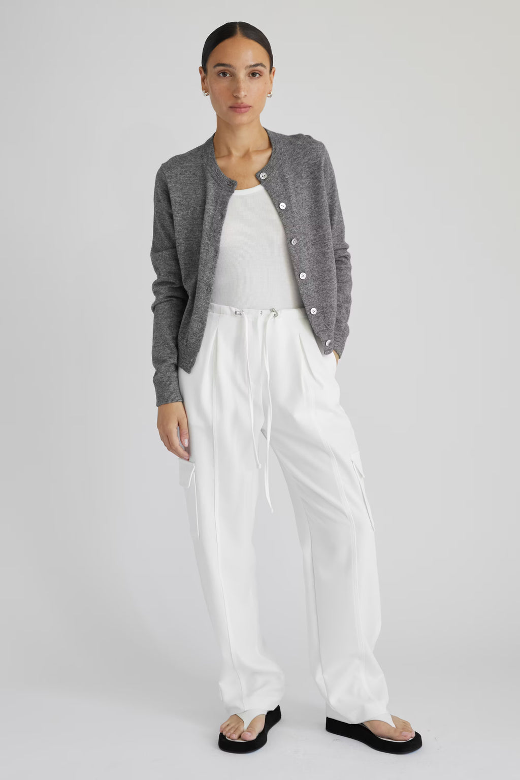 Woman wearing a gray cardigan, white top, and white pants on a light gray background