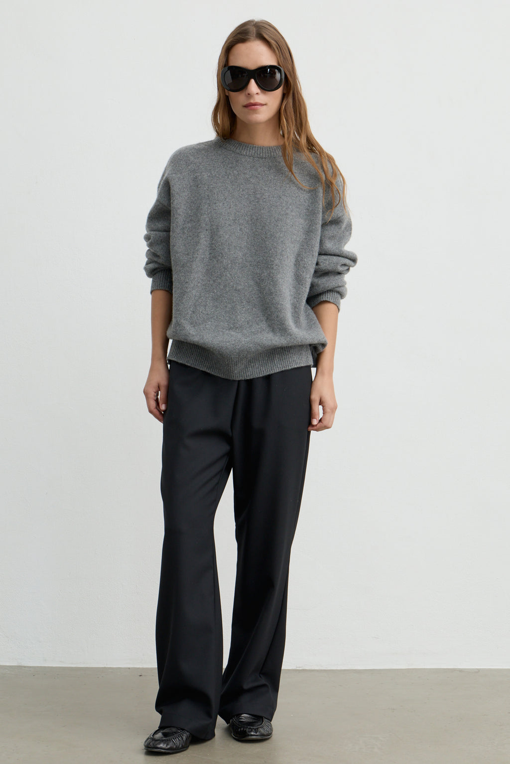 A woman modeling a gray knit crewneck sweater and black wide-leg trousers against a plain light gray studio background.