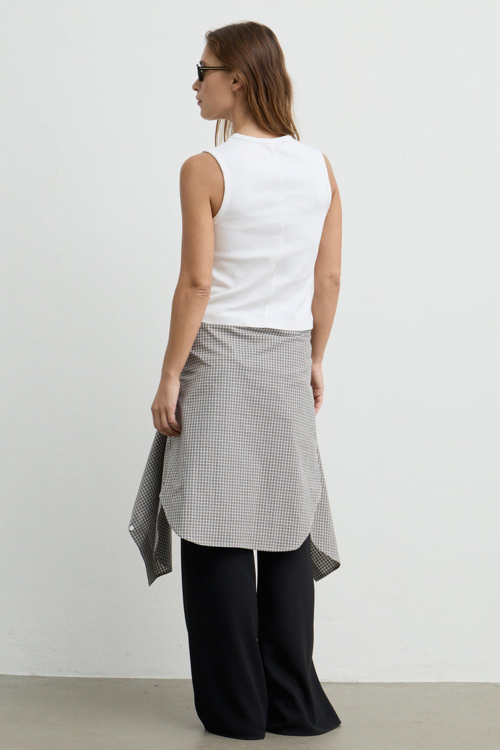 A woman shown from the back wearing a white sleeveless top, a gray and white gingham wrap skirt, and black wide-leg pants against a plain light gray studio background.