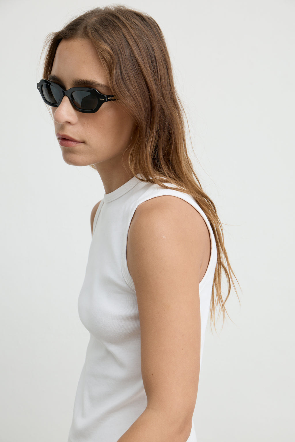 A woman in profile wearing black cat-eye sunglasses and a white sleeveless top, photographed against a plain light gray studio background.
