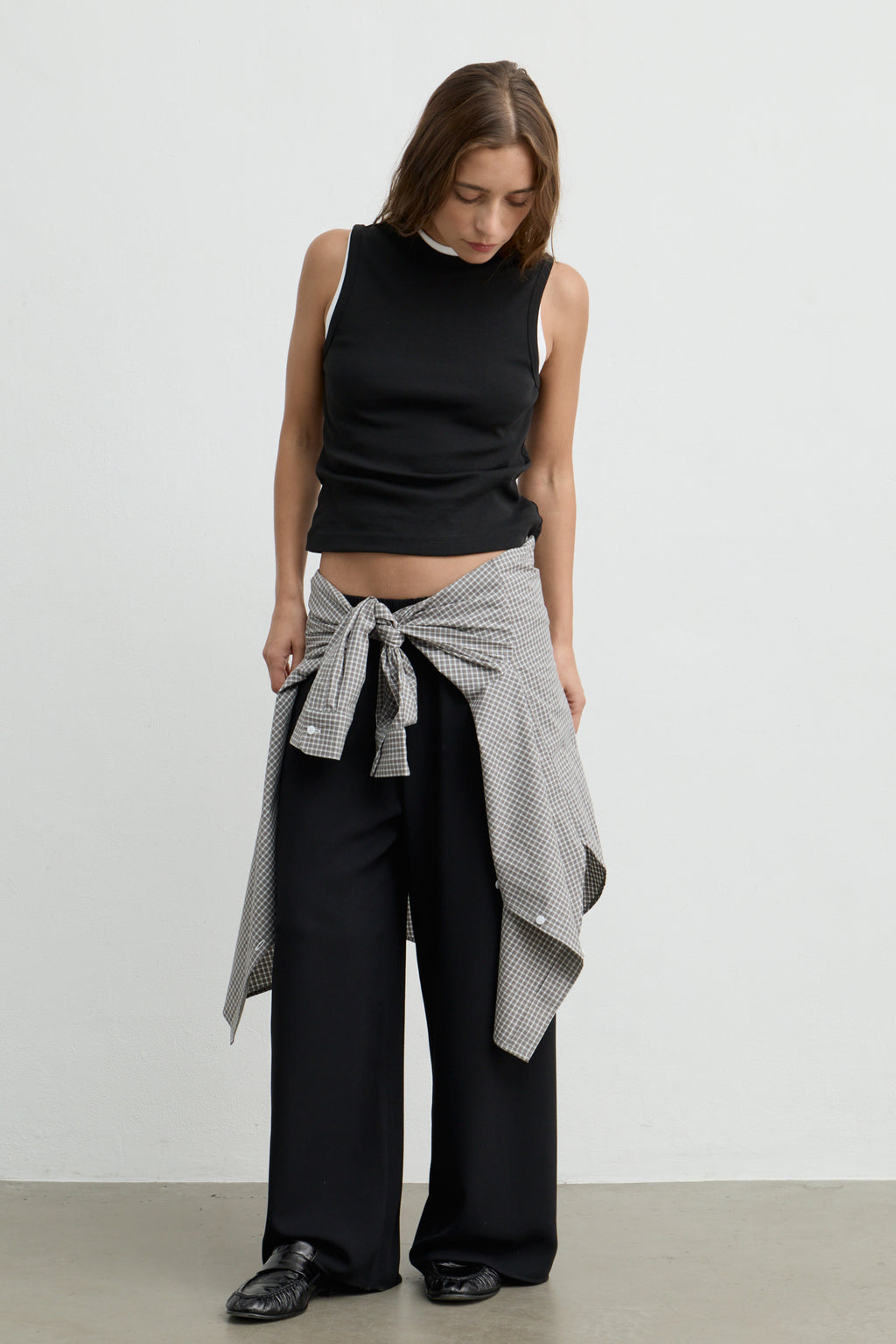 A woman stands against a plain light gray wall wearing a black sleeveless mock-neck top, black wide-leg pants, and a black-and-white patterned shirt tied around her waist.