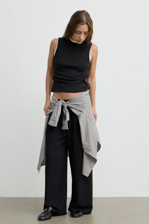 A woman stands against a plain light gray wall wearing a black sleeveless mock-neck top, black wide-leg pants, and a black-and-white patterned shirt tied around her waist.