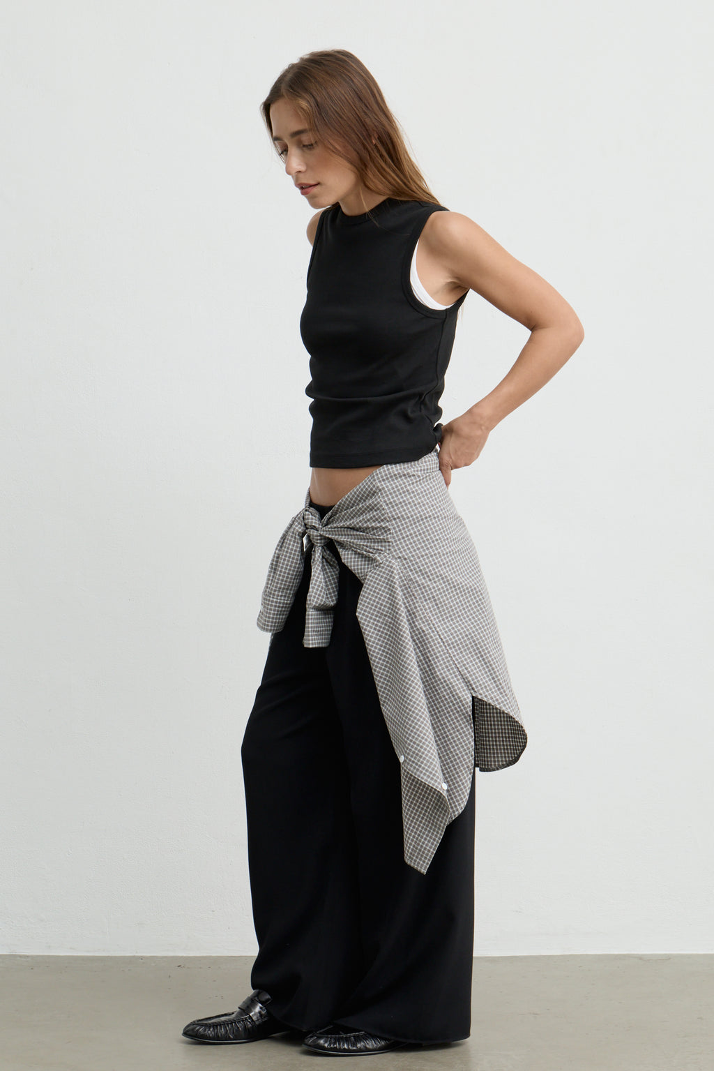 A woman stands in profile wearing a black sleeveless tank top, loose black wide-leg pants, and a black-and-white patterned shirt tied around her waist against a plain light gray studio background.