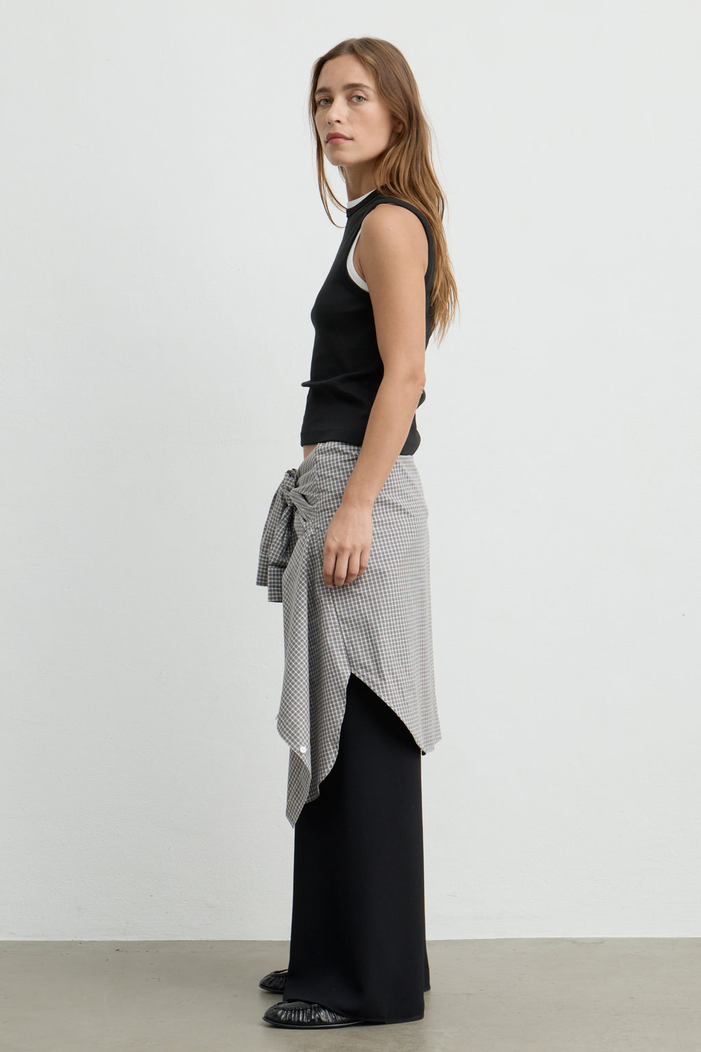 A woman stands in profile wearing a black sleeveless top, wide-leg black pants, and a gray-and-white patterned wrap shirt tied at the waist, photographed against a plain light gray studio background.