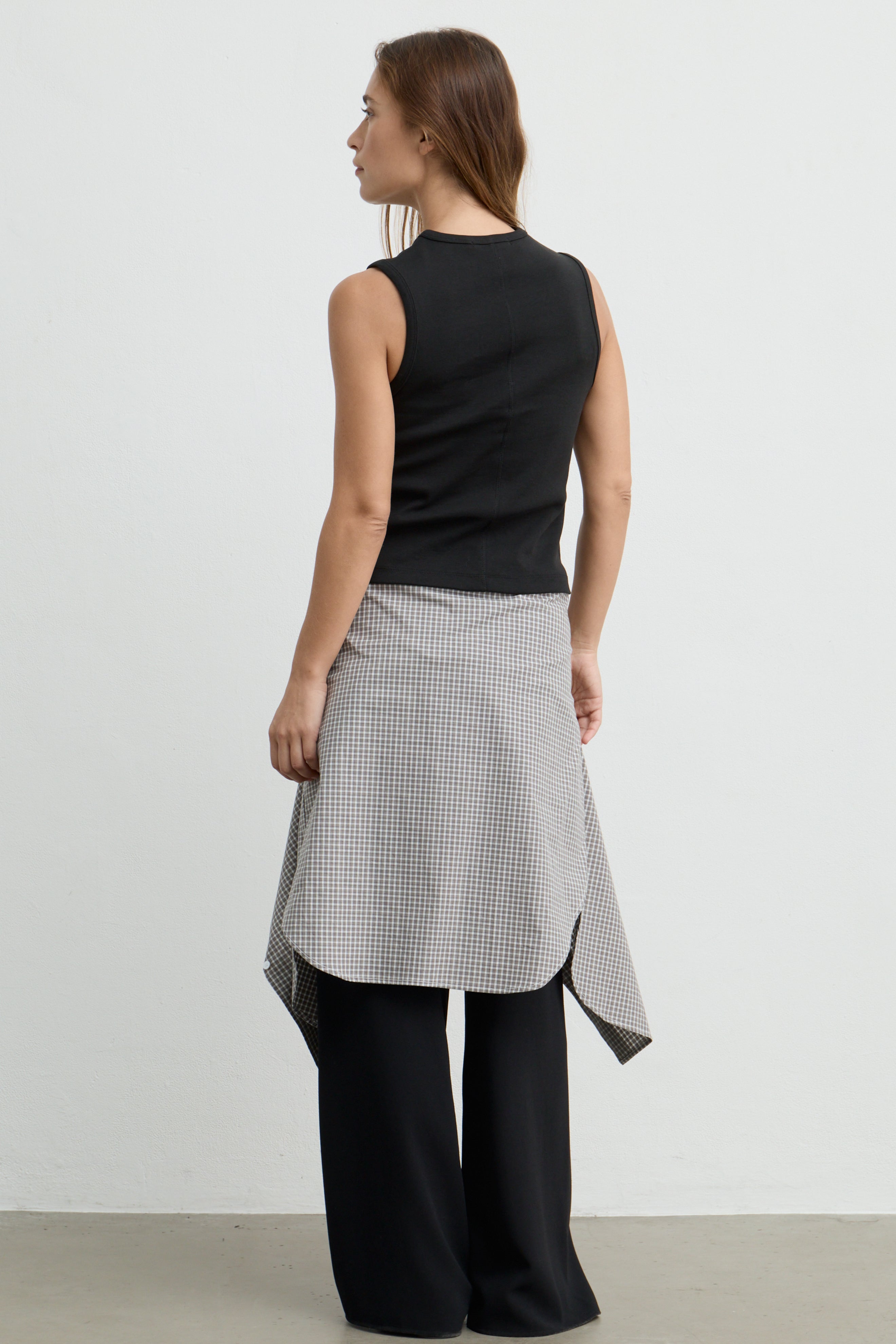 A woman shown from the back wearing a sleeveless black top, a black-and-white gingham wrap shirt, and black wide-leg pants against a plain light gray studio background.