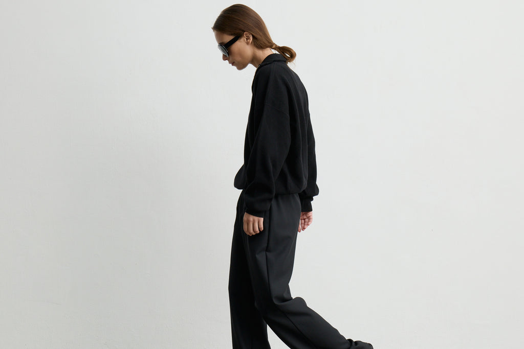 A model in side profile wearing a black long-sleeve sweatshirt and matching black wide-leg trousers, photographed against a plain light gray studio background.