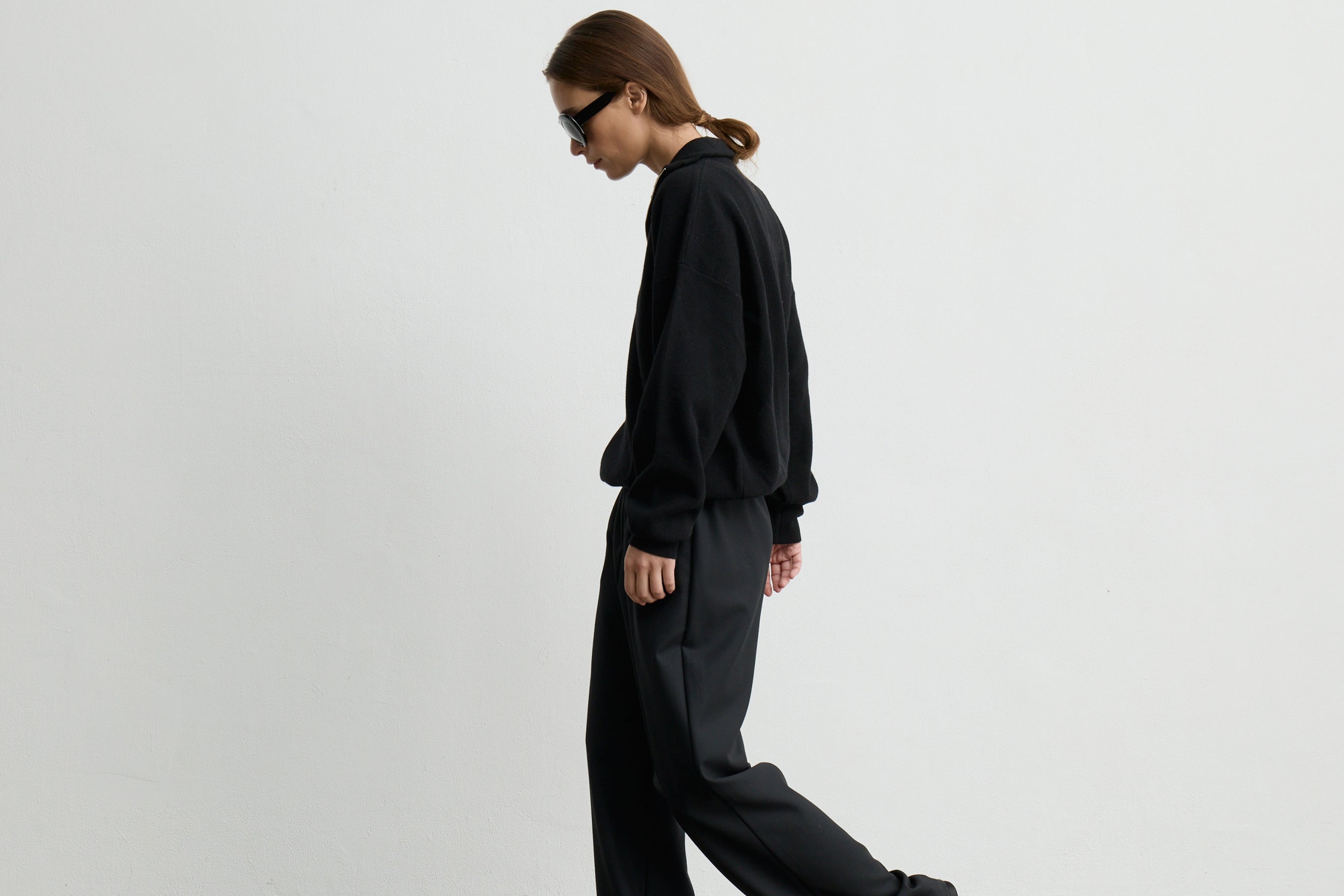 A model in side profile wearing a black long-sleeve sweatshirt and matching black wide-leg trousers, photographed against a plain light gray studio background.