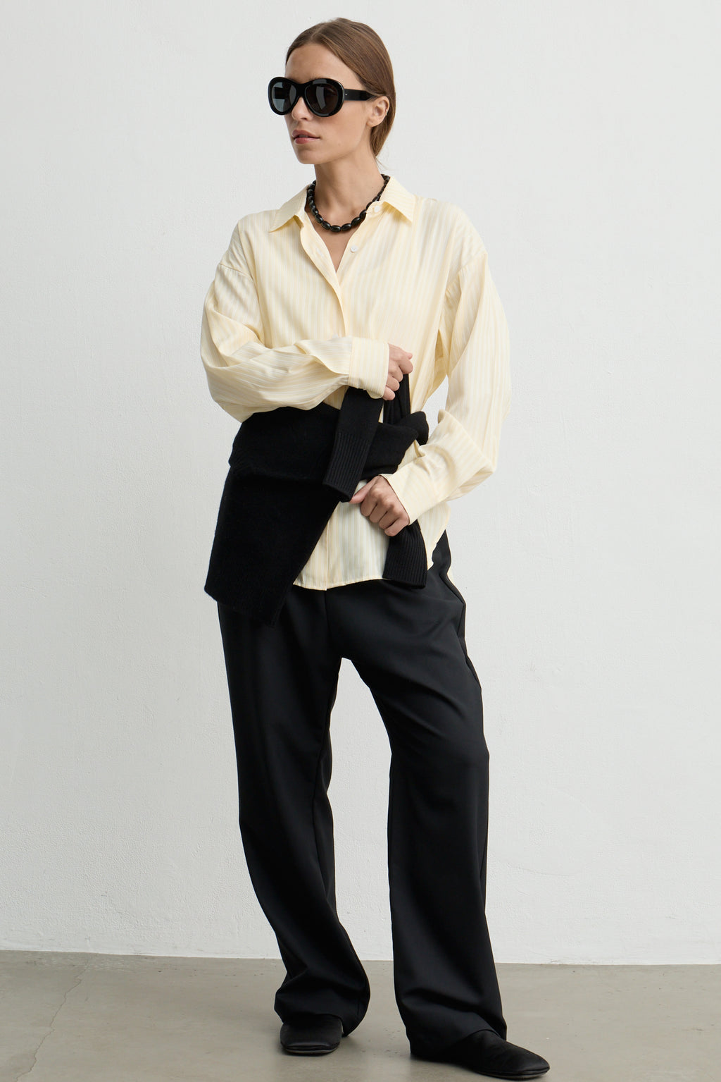 A model stands against a plain light gray wall wearing a yellow vertically striped button-down shirt, black wide-leg trousers, black pointed shoes, black sunglasses, and a black beaded necklace.