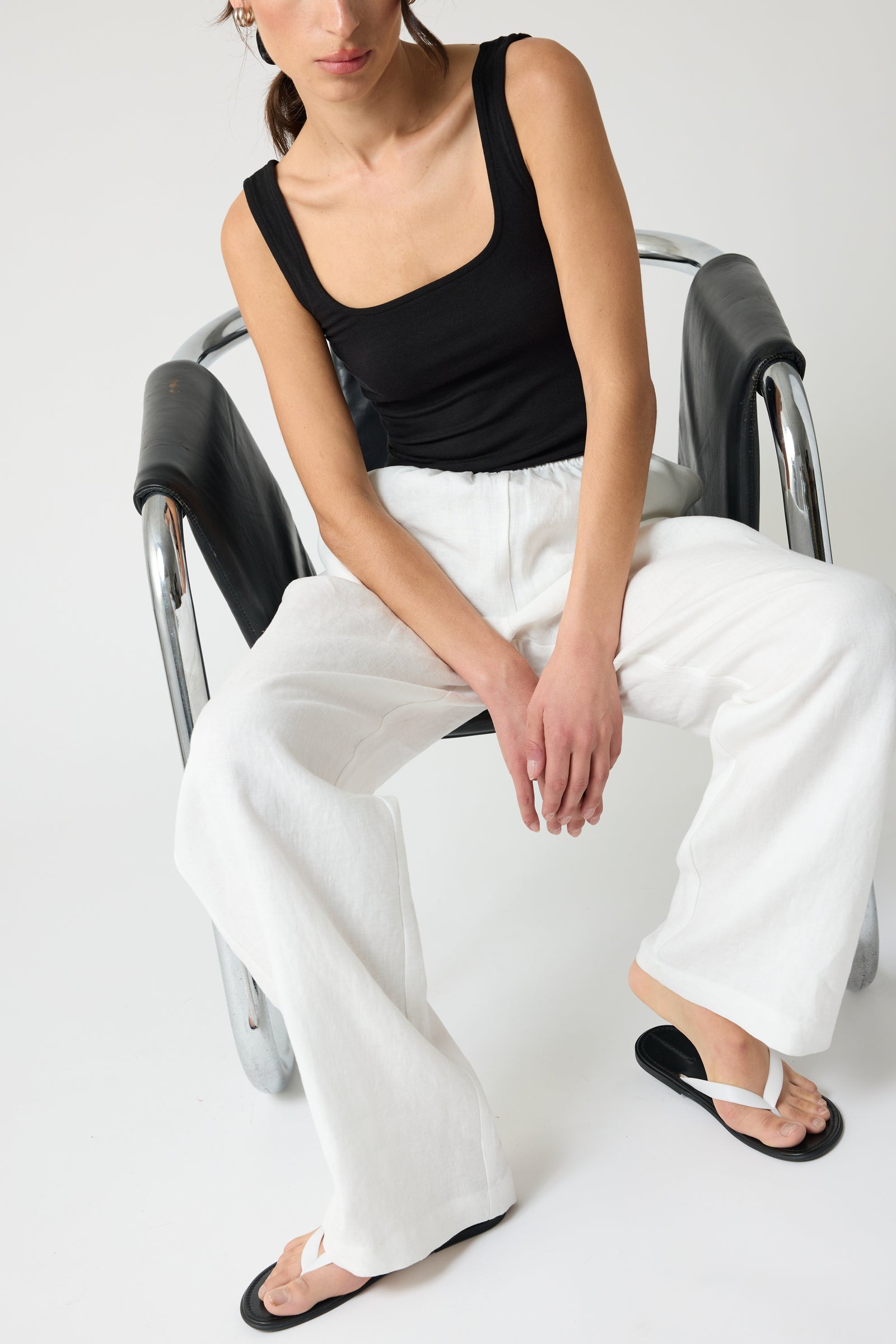 Woman sitting in a chair wearing a black top and white linen pants on a plain background