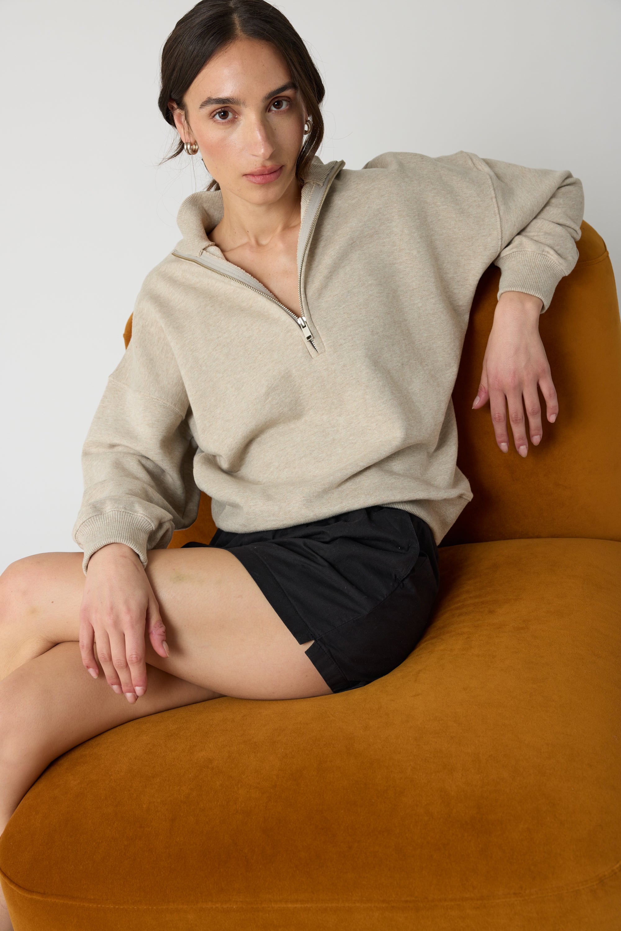 Woman wearing a beige zip up sweater and black shorts sitting on an orange couch against a white background