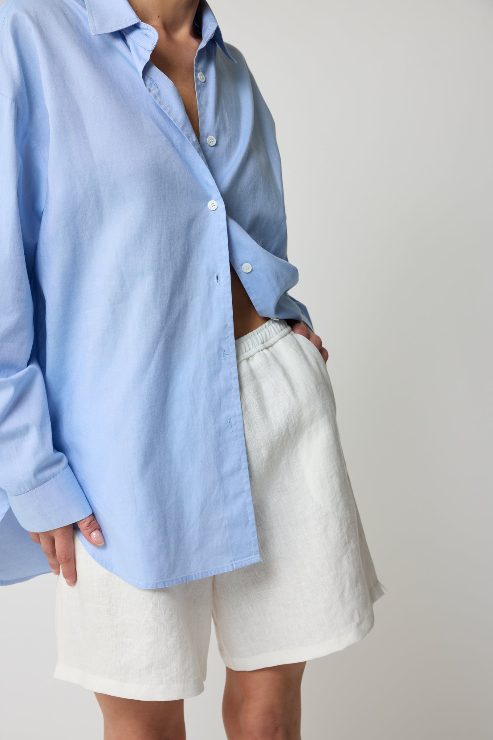 Woman wearing a light cotton button up blue shirt and white linen shorts on a plain background