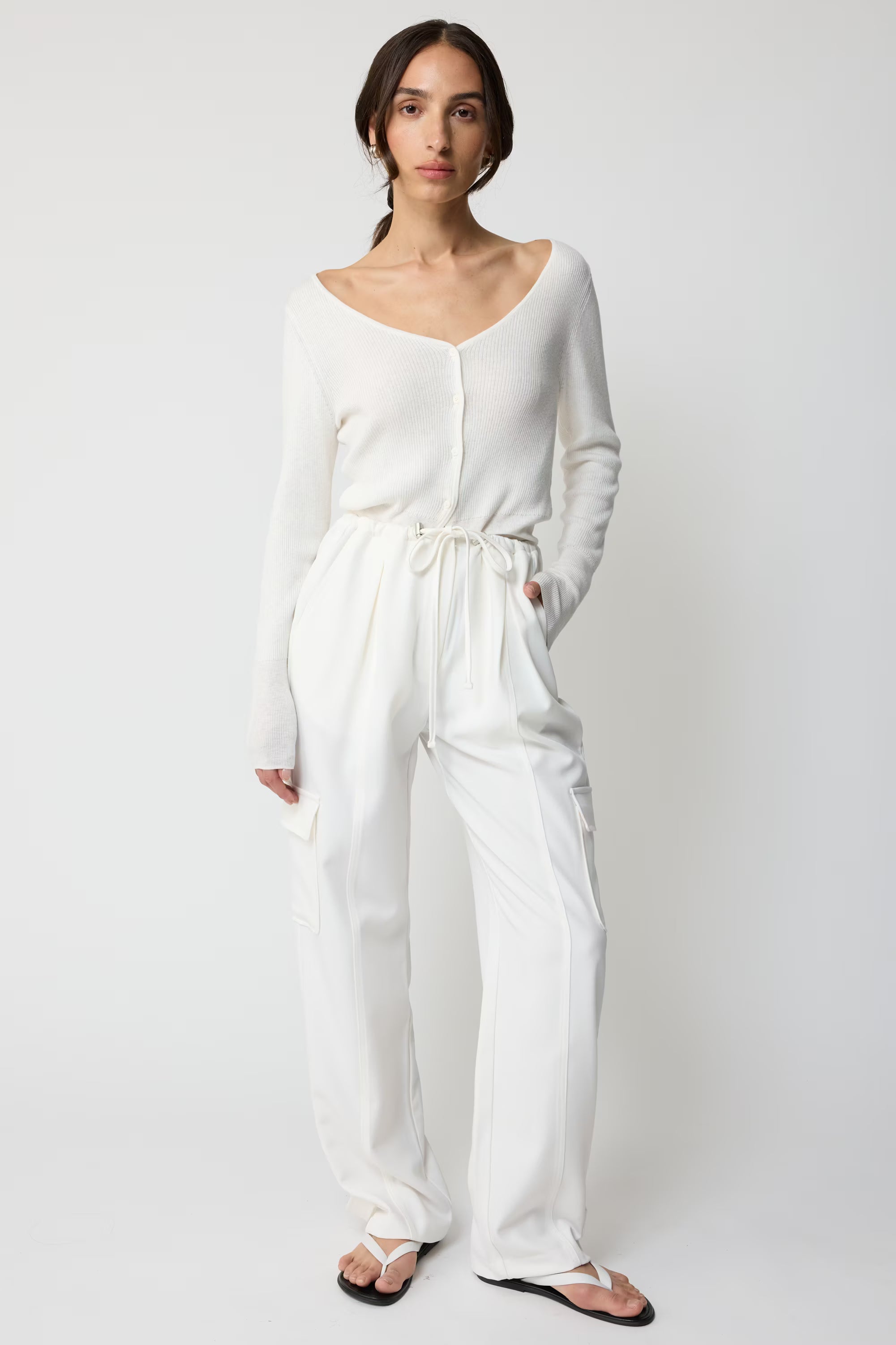 Woman wearing a white cardigan and white cargo pants on a plain background