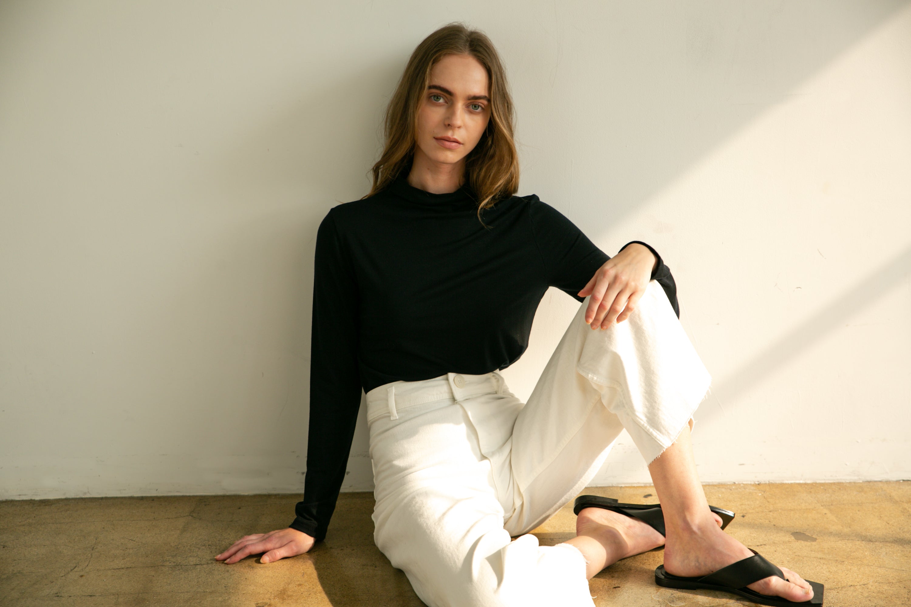 Woman sitting on the floor wearing a black top and white pants with a neutral background