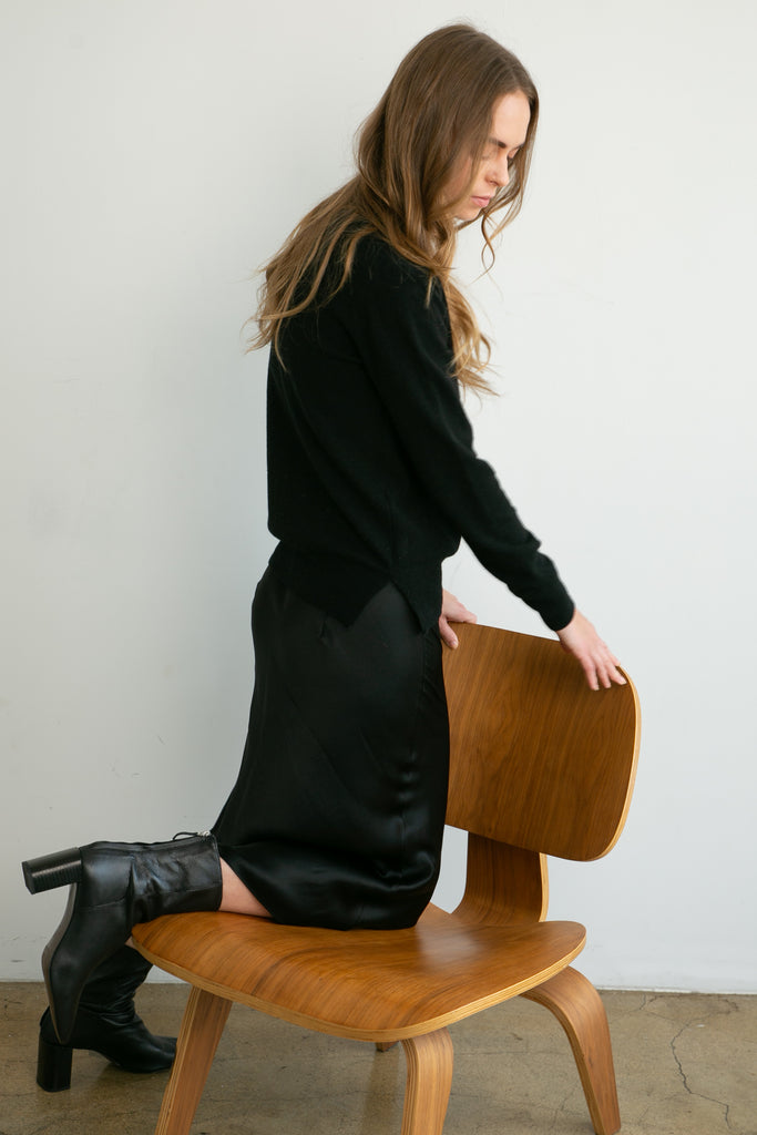 Woman in a black wool sweater over a silk black skirt and standing next to a wooden chair against a white wall.