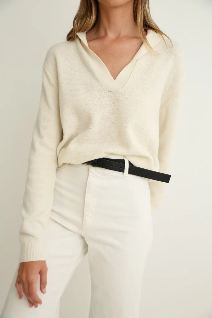 Woman wearing a cream sweater and white pants with a black belt against a plain background