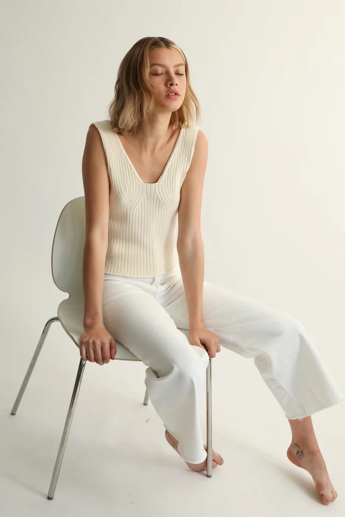 Woman sitting on a chair wearing a cream sleeveless top and white pants against a plain background