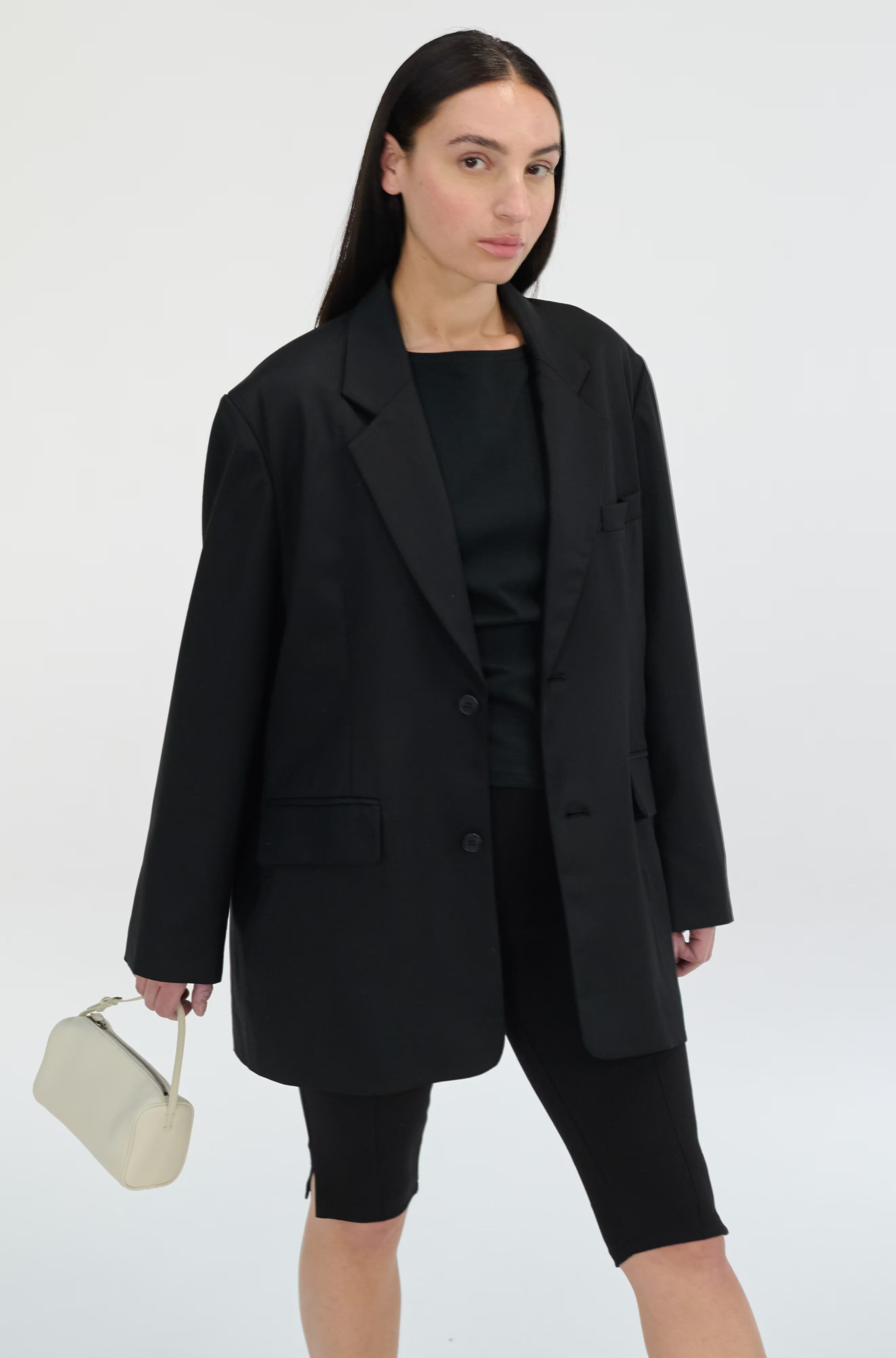 Woman wearing a black blazer and black outfit holding a beige handbag on a white background