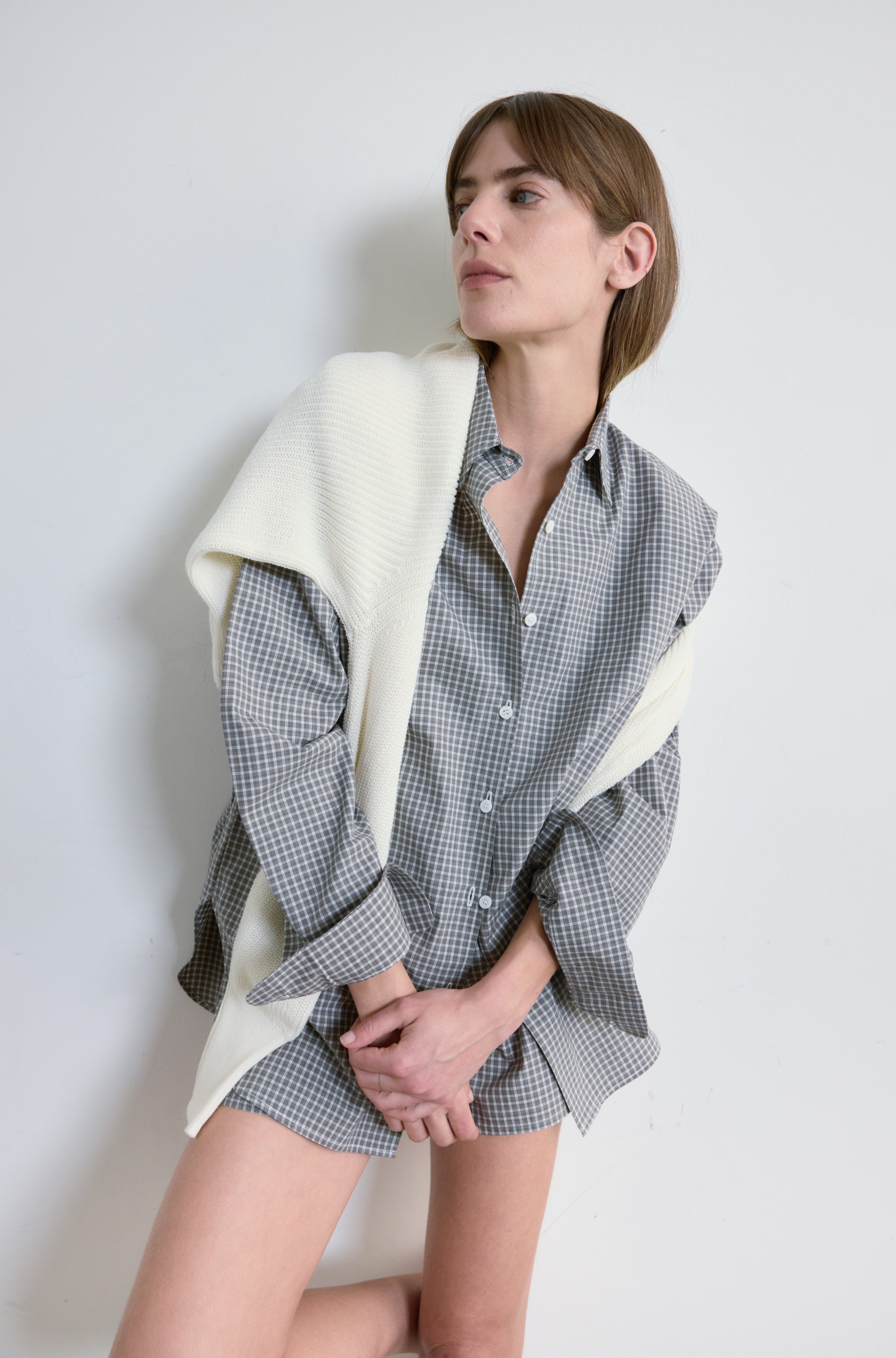 Woman wearing a checkered shirt and shorts with a white blanket draped over her shoulders against a plain background