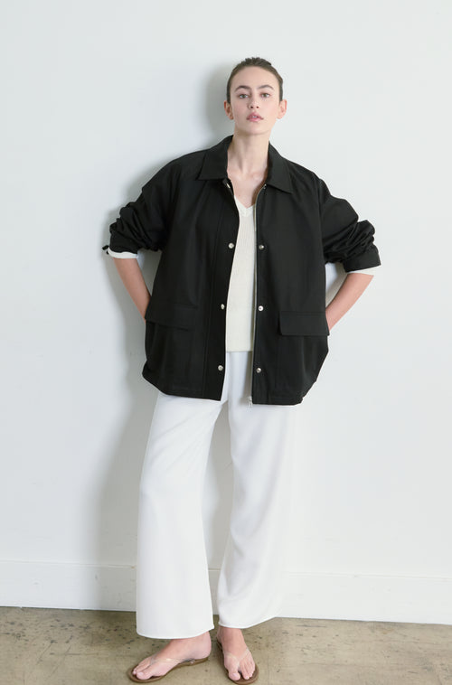 Person wearing a black jacket and white pants against a white wall