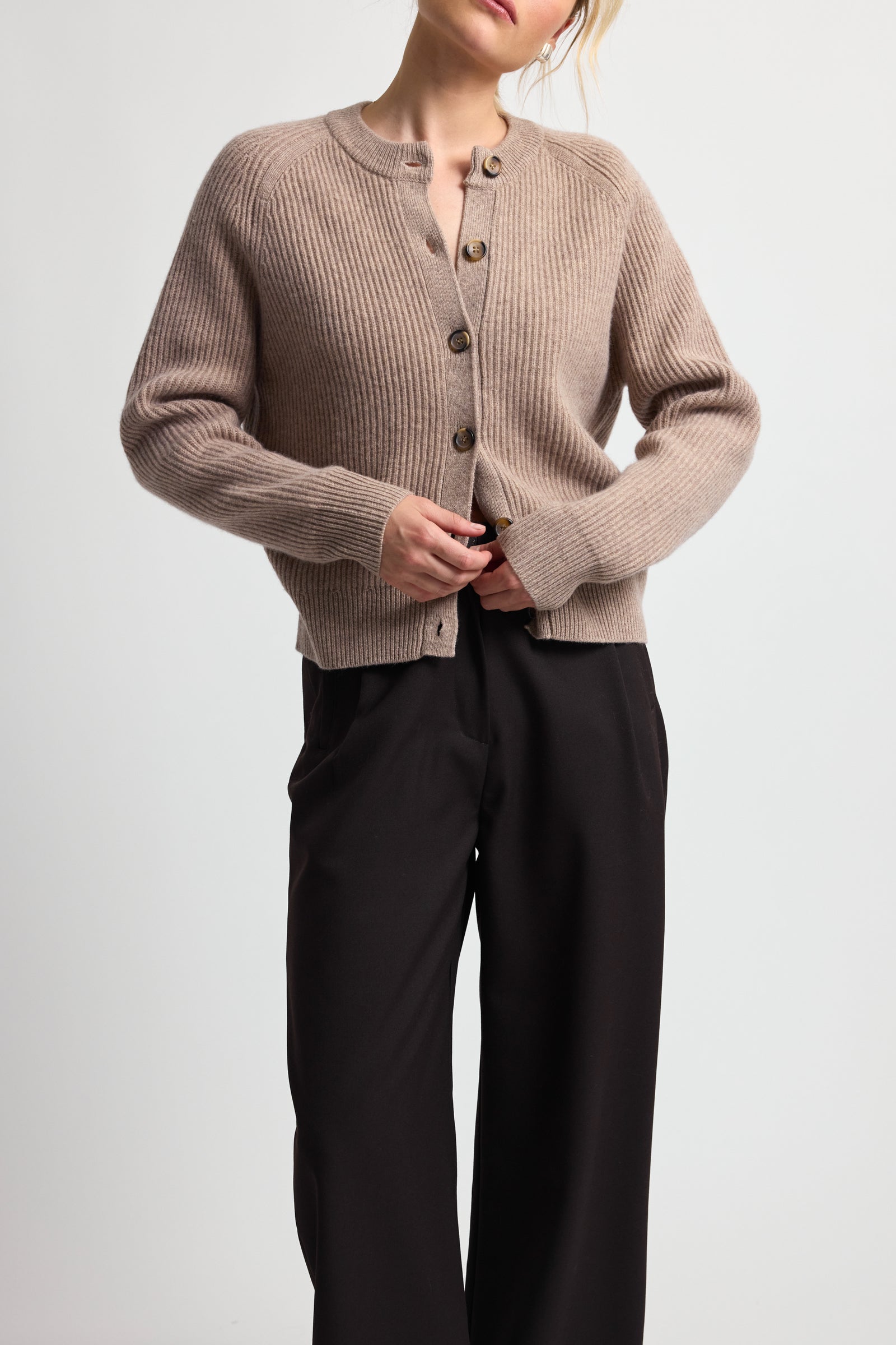 Woman wearing a beige button up cardigan and dark brown trousers on a light gray background