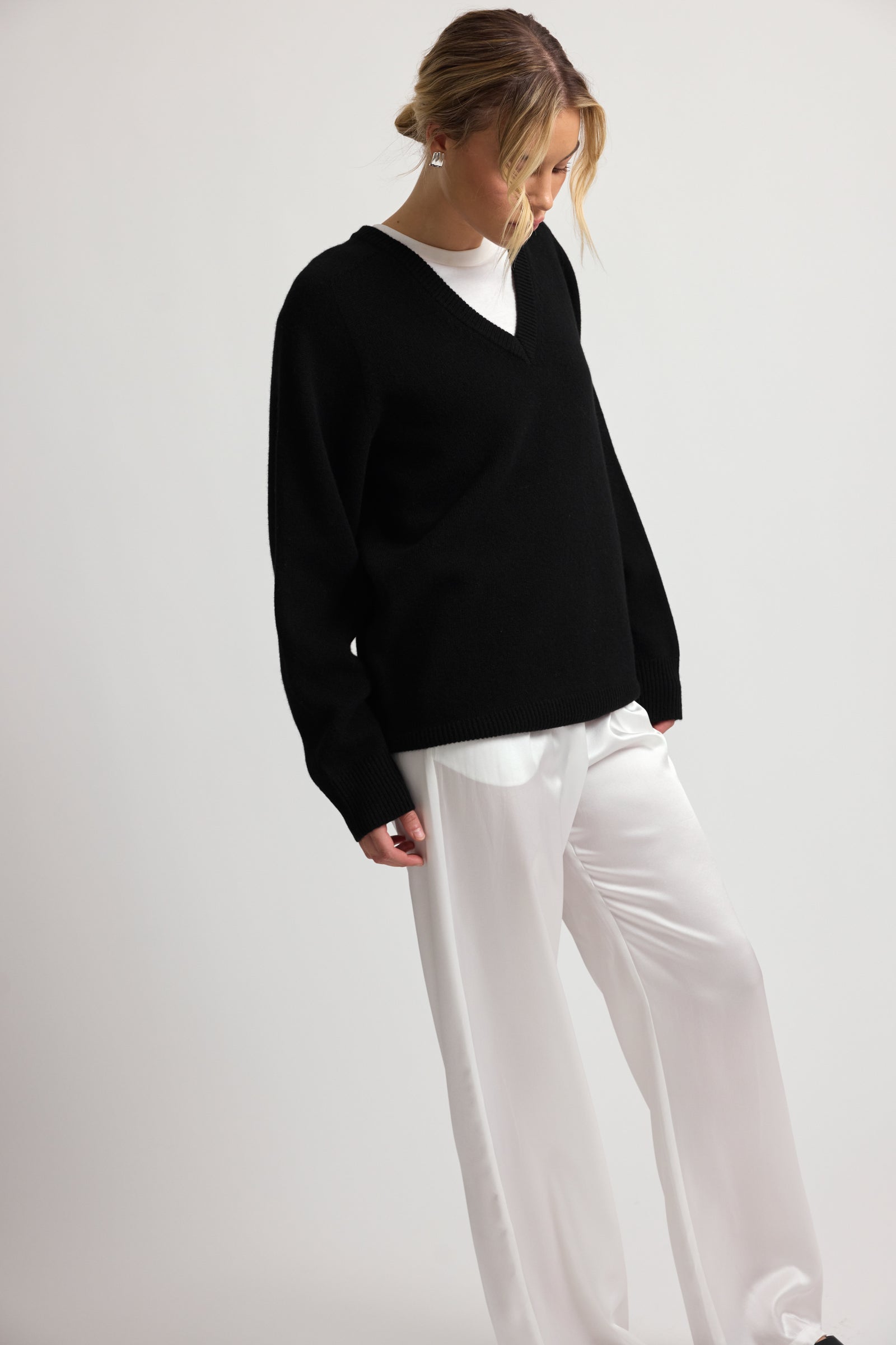 Woman wearing a v neck wool black sweater and silk white pants on a plain background