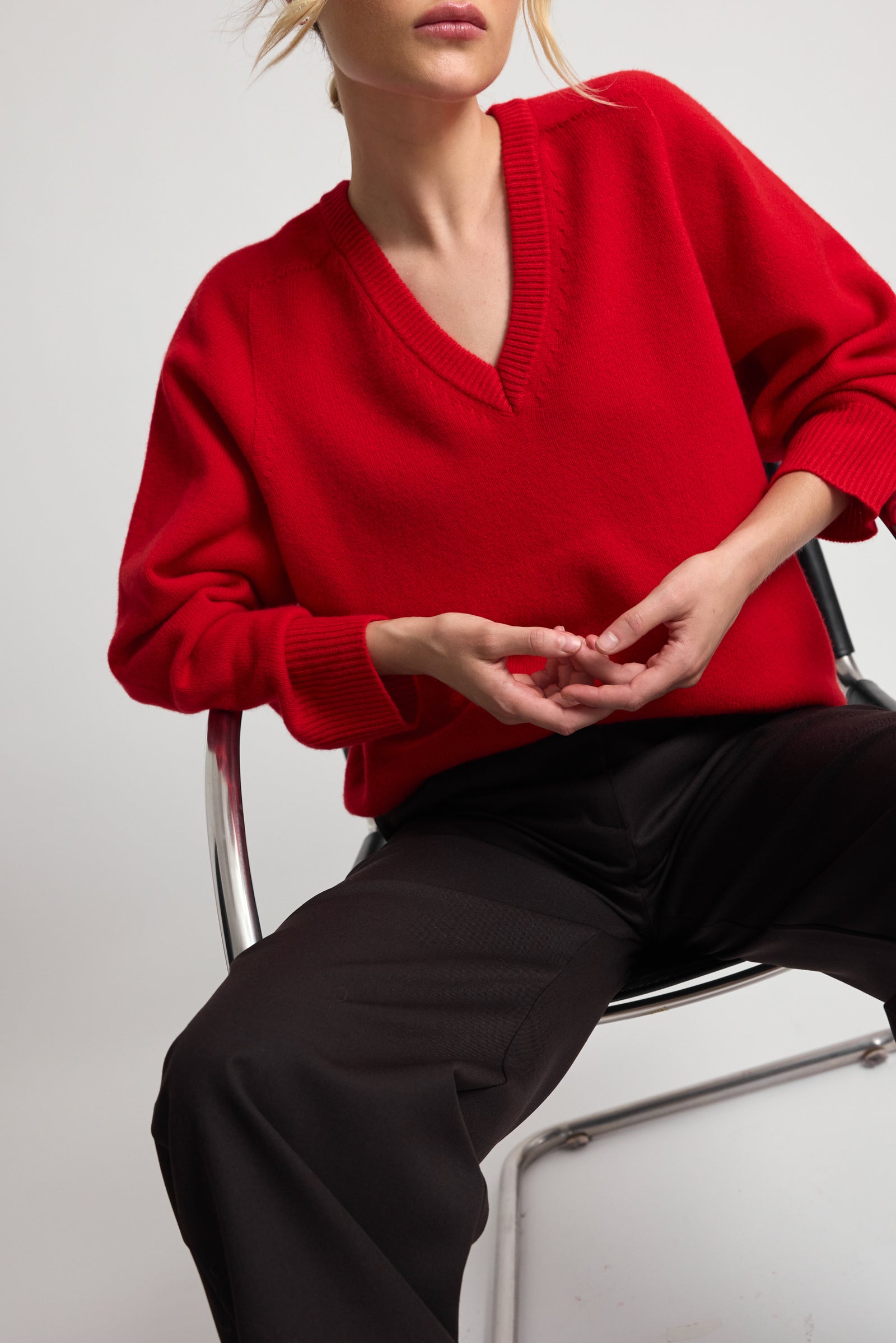 Woman wearing a red v-neck sweater and dark brown pants sitting on a white chair against a light gray background