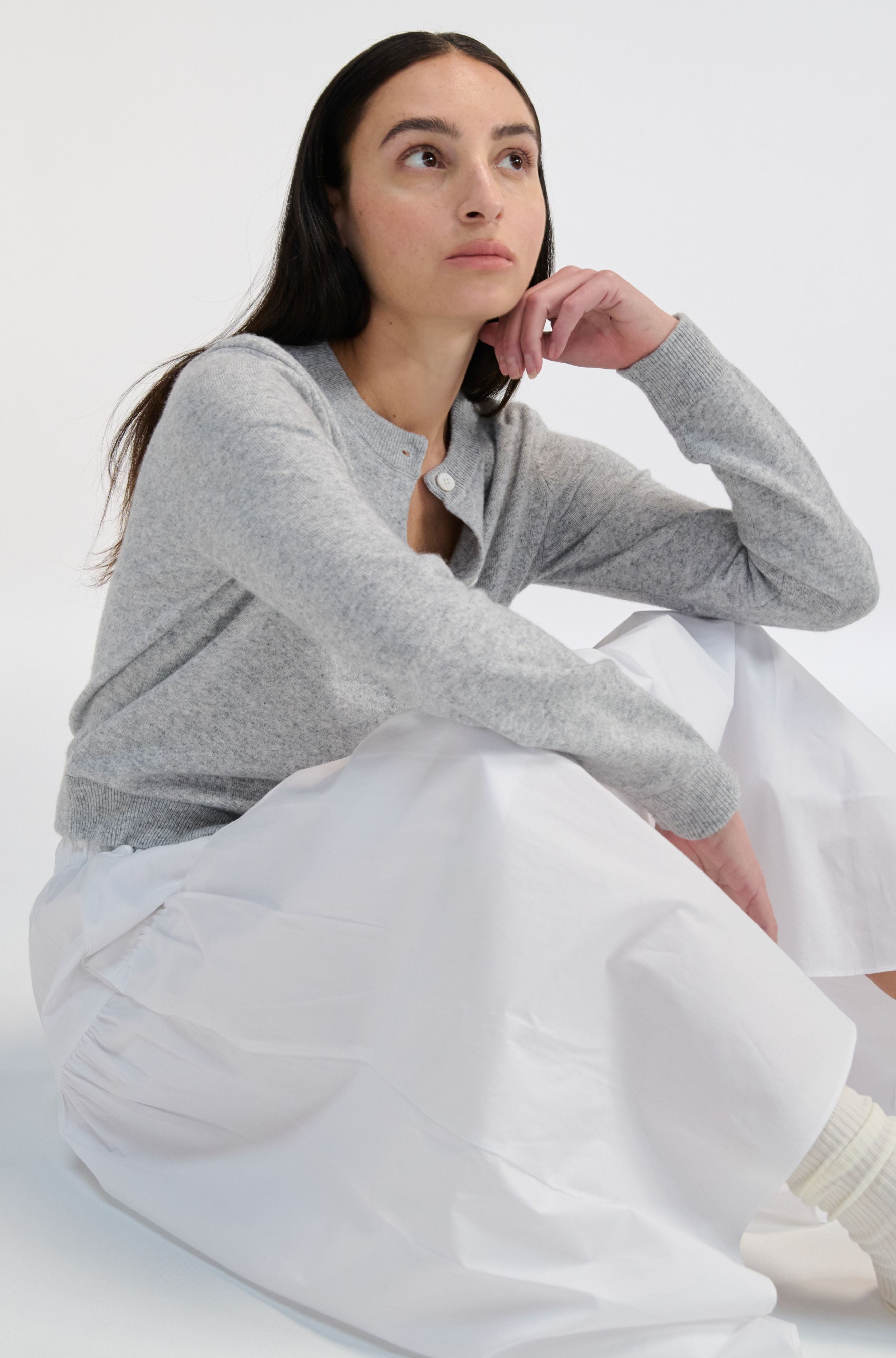Woman wearing a gray sweater and white skirt sitting on a white background