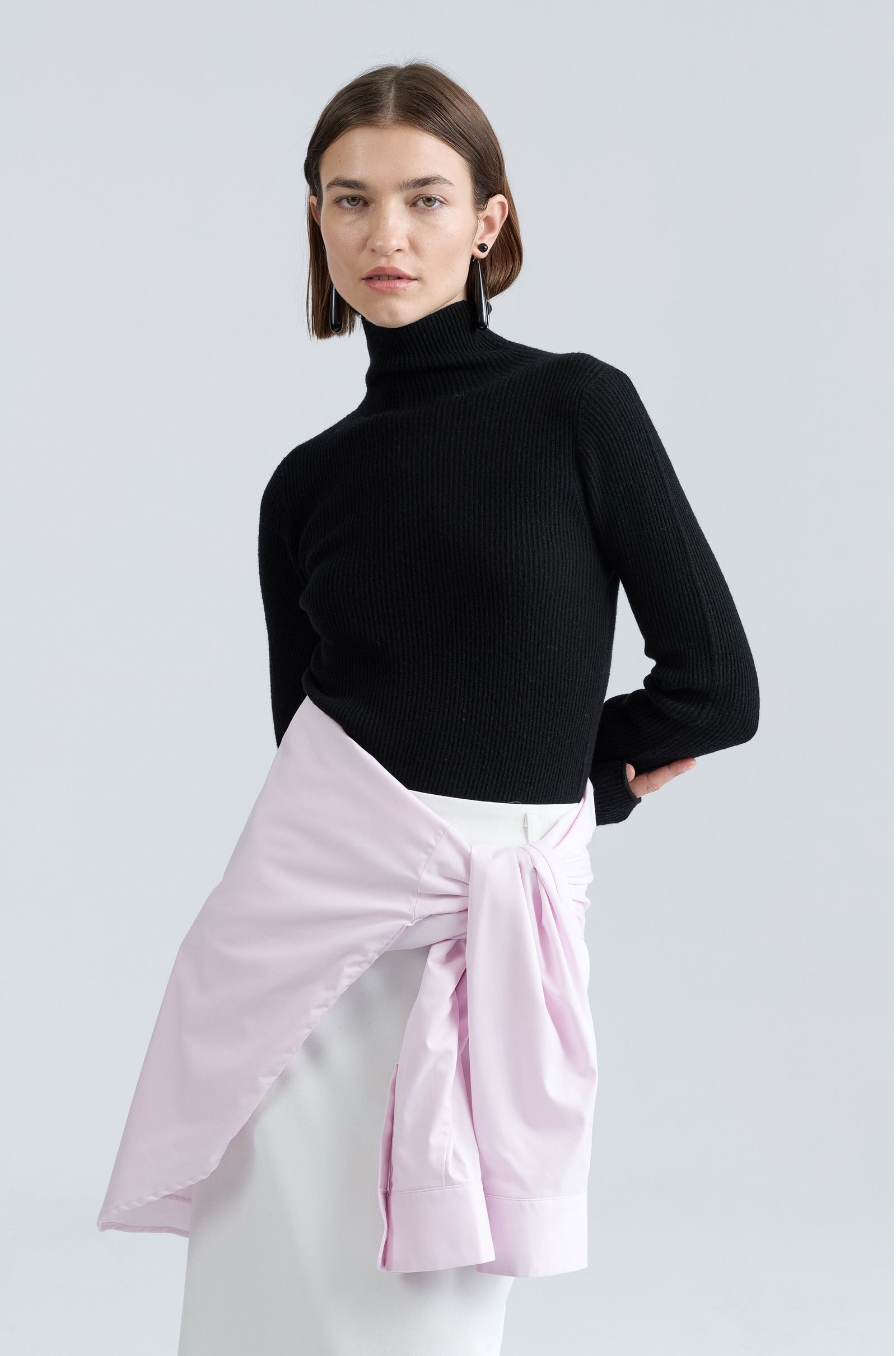Person wearing a black turtleneck and light pink wrap over white pants on a plain background