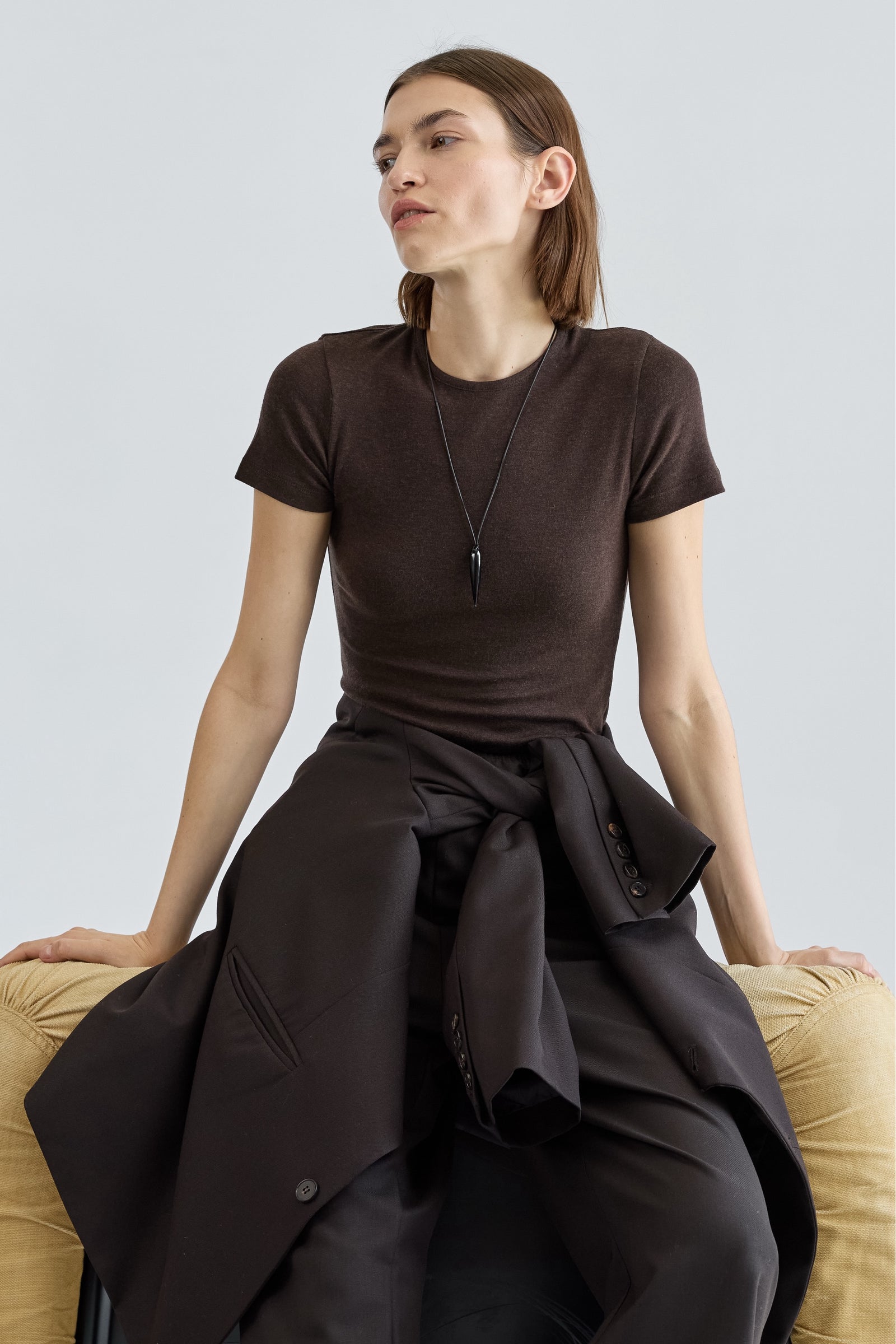 Woman wearing a brown t-shirt and dark coat sitting on a beige couch against a light gray background