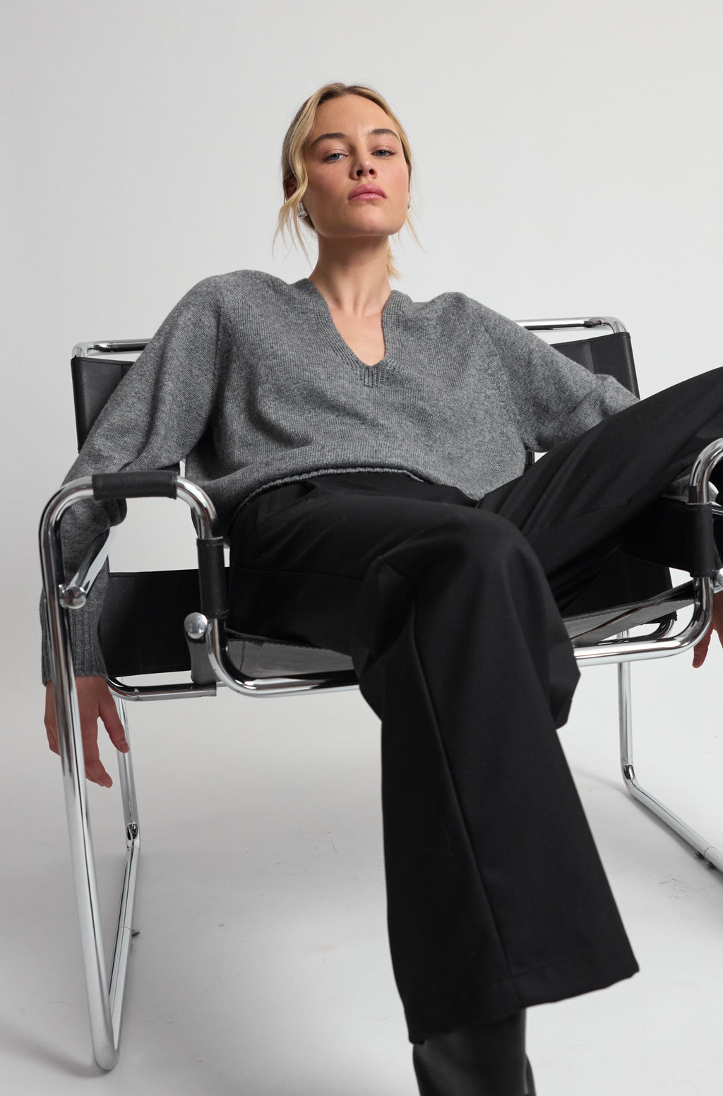 Woman sitting on a modern chair wearing a gray sweater and black pants against a white background