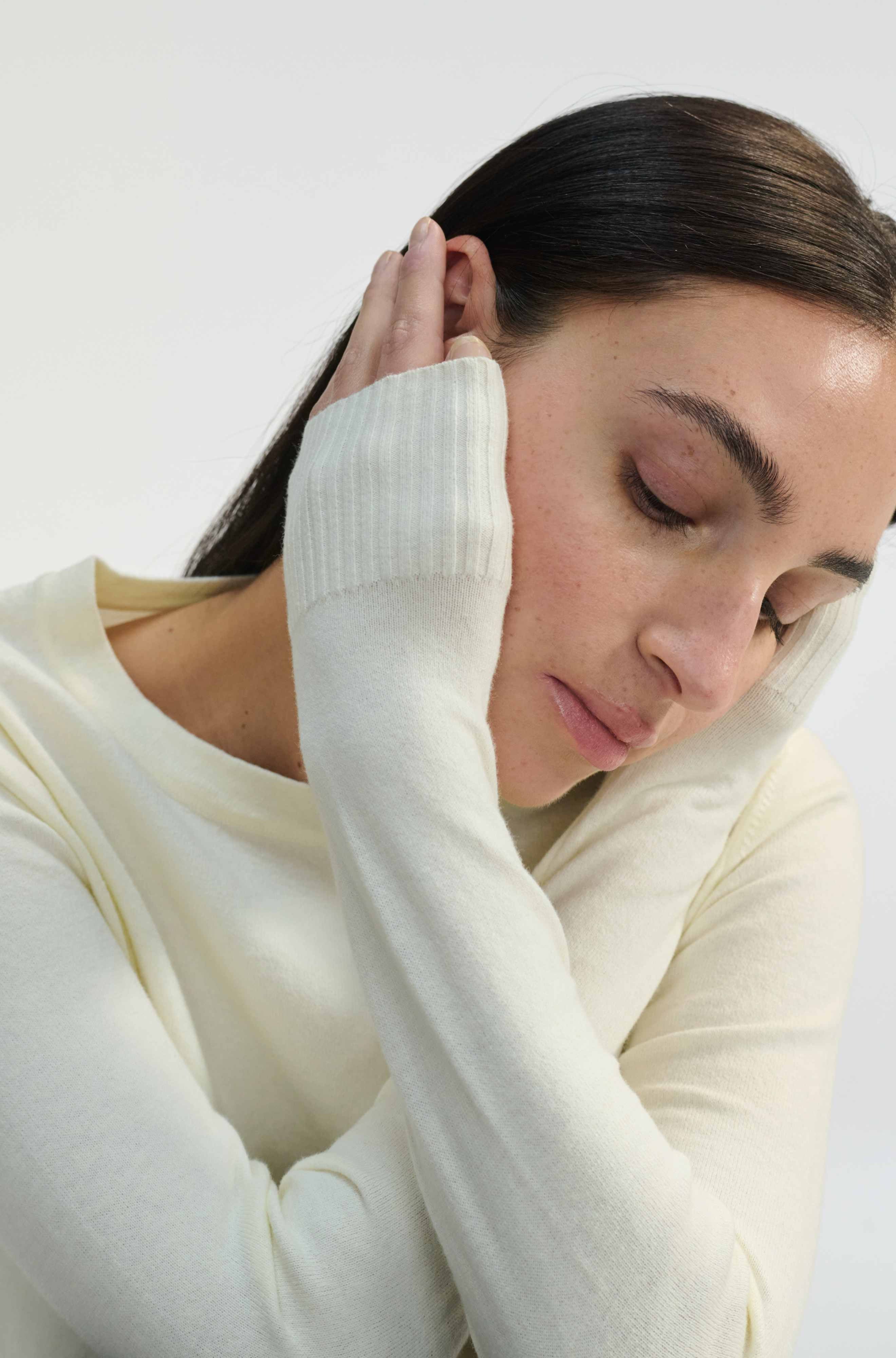 Woman in a white sweater with her hand on her head against a plain background