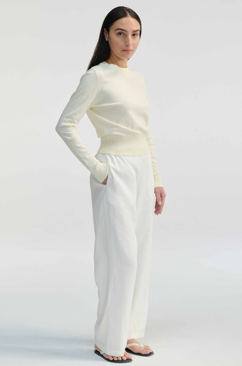 Woman wearing a cream long-sleeve top and white pants on a light gray background