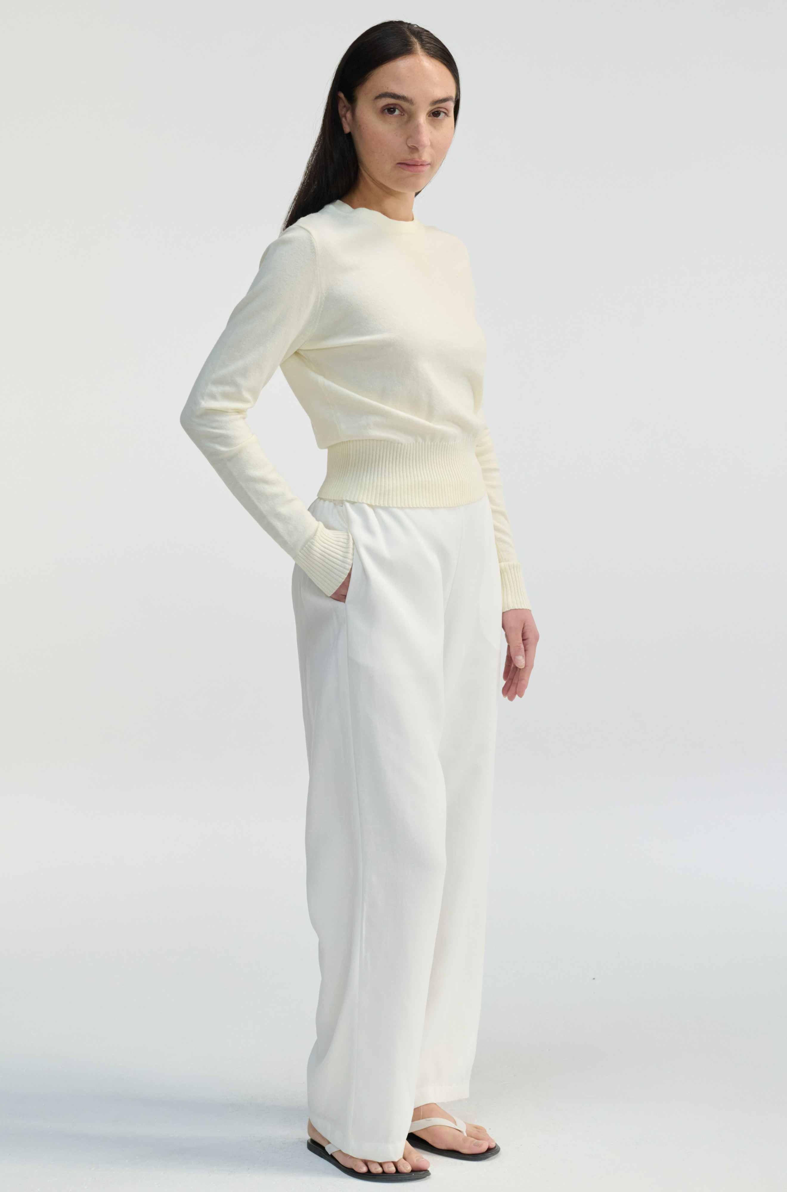 Woman wearing a cream long-sleeve top and white pants on a light gray background