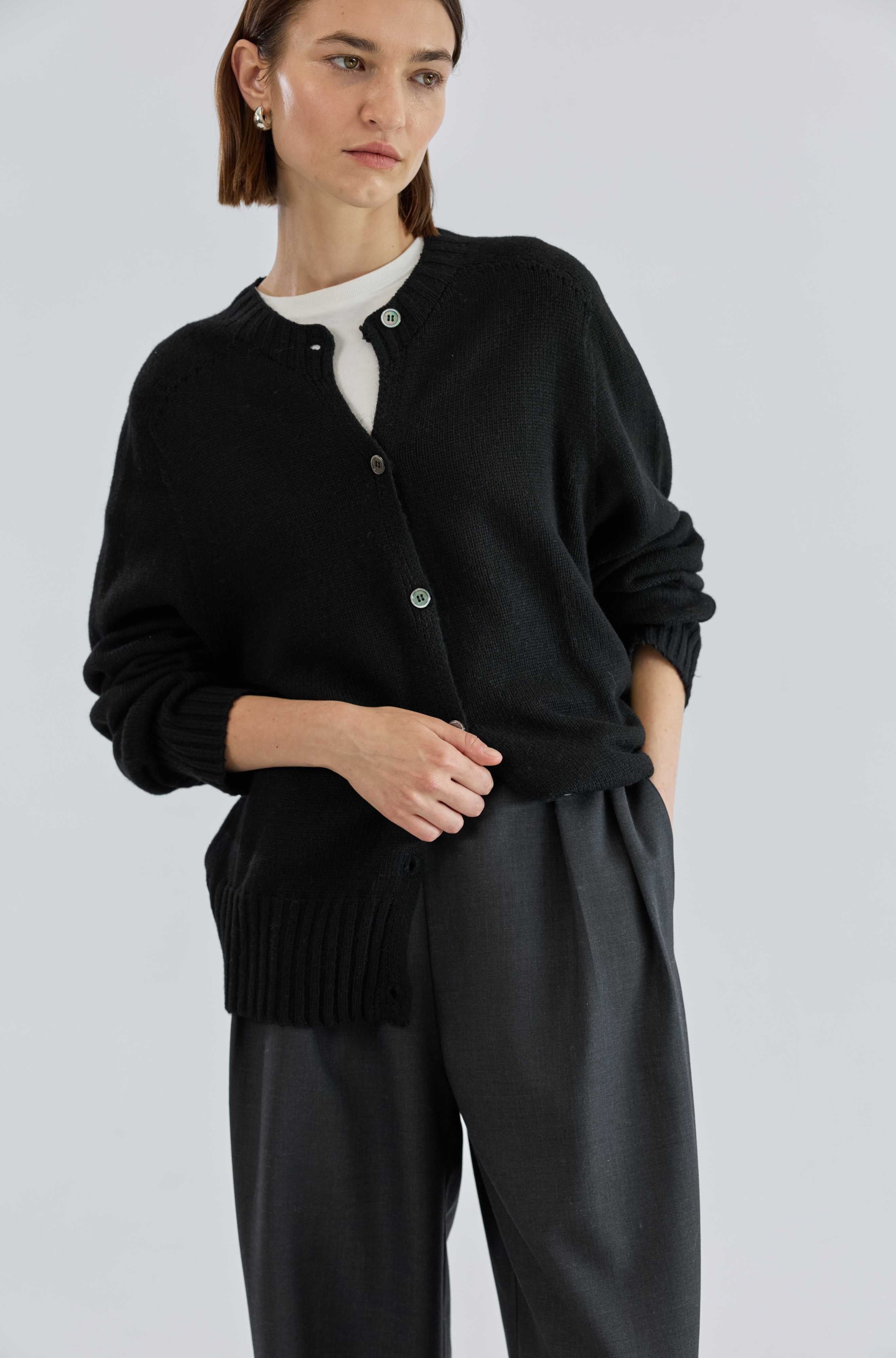 Woman wearing a black cardigan over a white shirt with gray pants on a light gray background