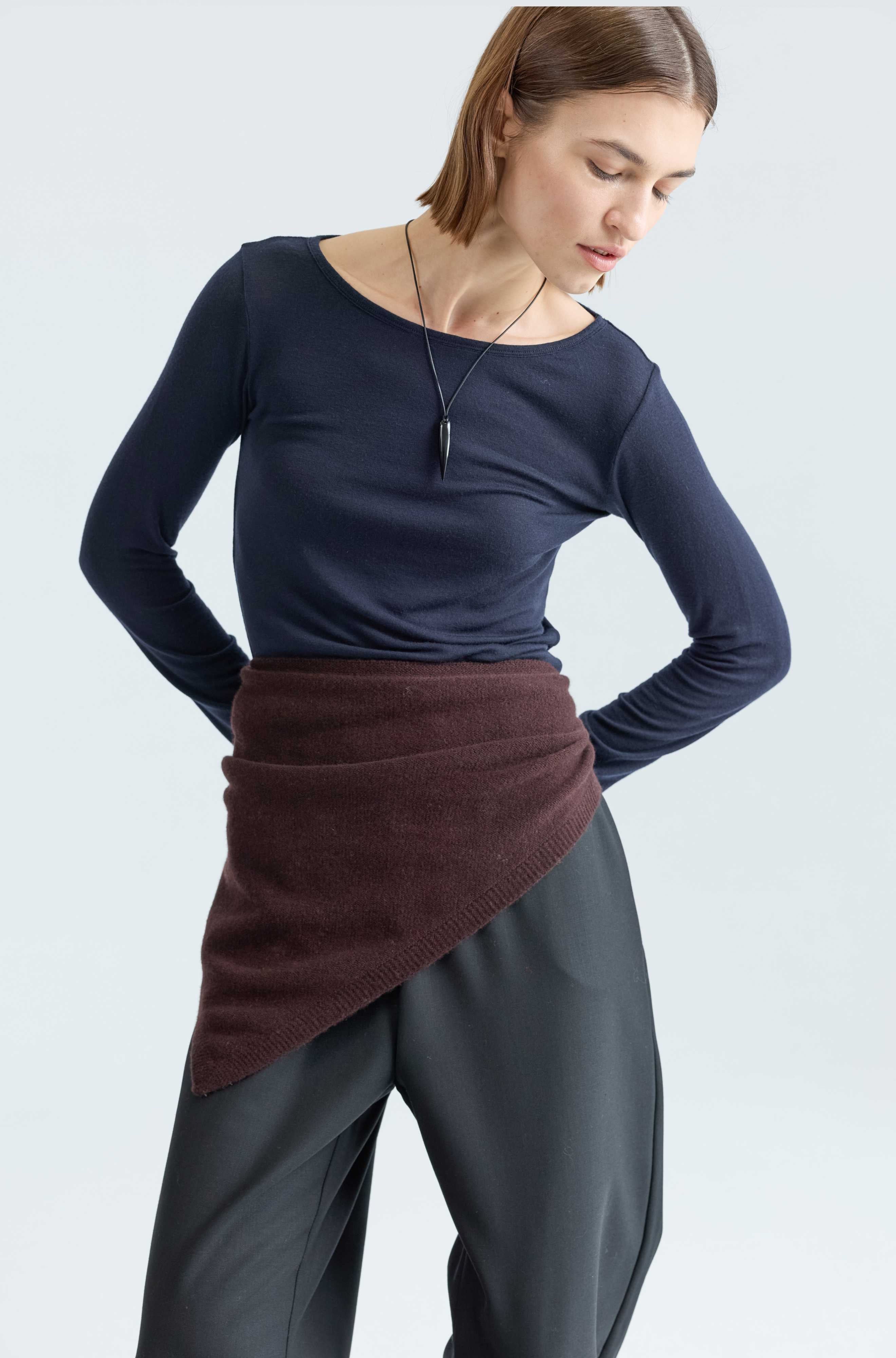 Woman wearing a navy long-sleeve top and dark pants with a brown wrap around her waist on a light gray background