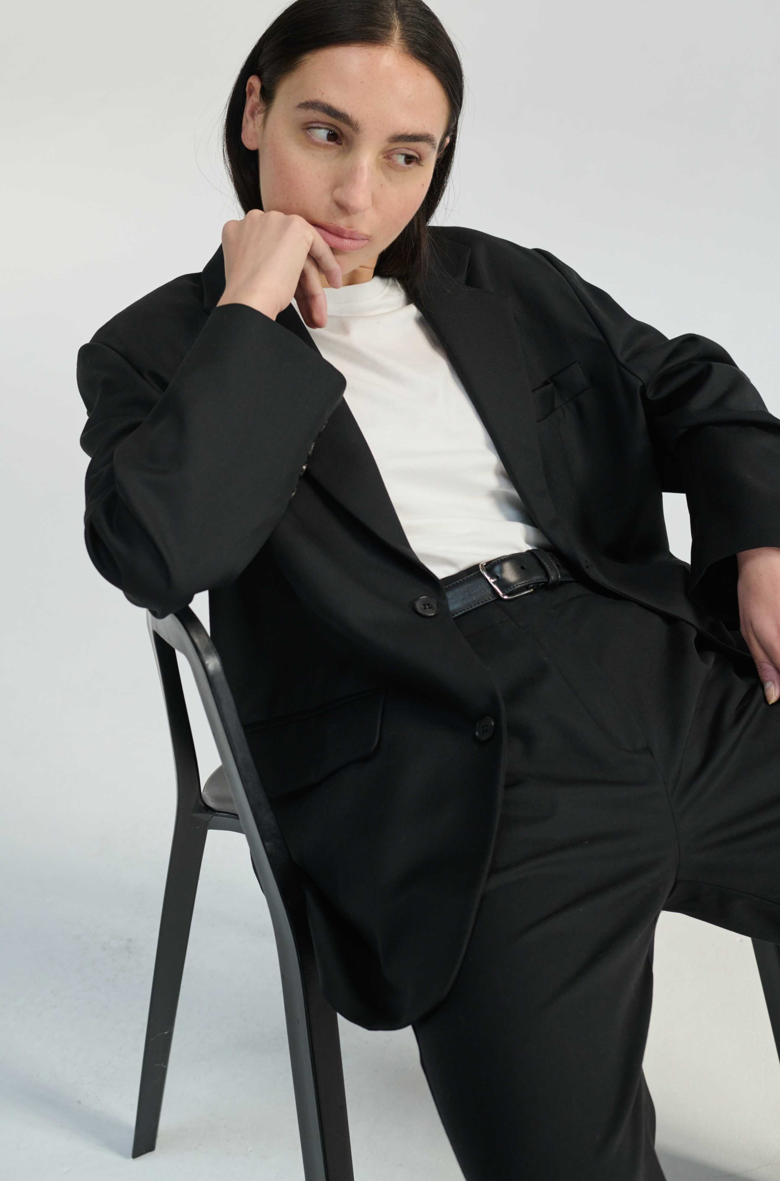 Person wearing a black suit and white turtleneck sitting on a chair against a plain background