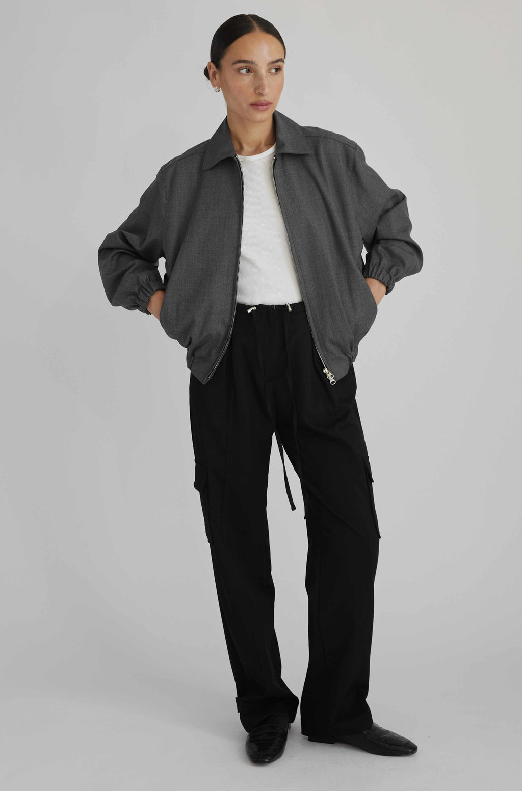 Person wearing a gray jacket, white shirt, black pants, and black shoes on a light gray background