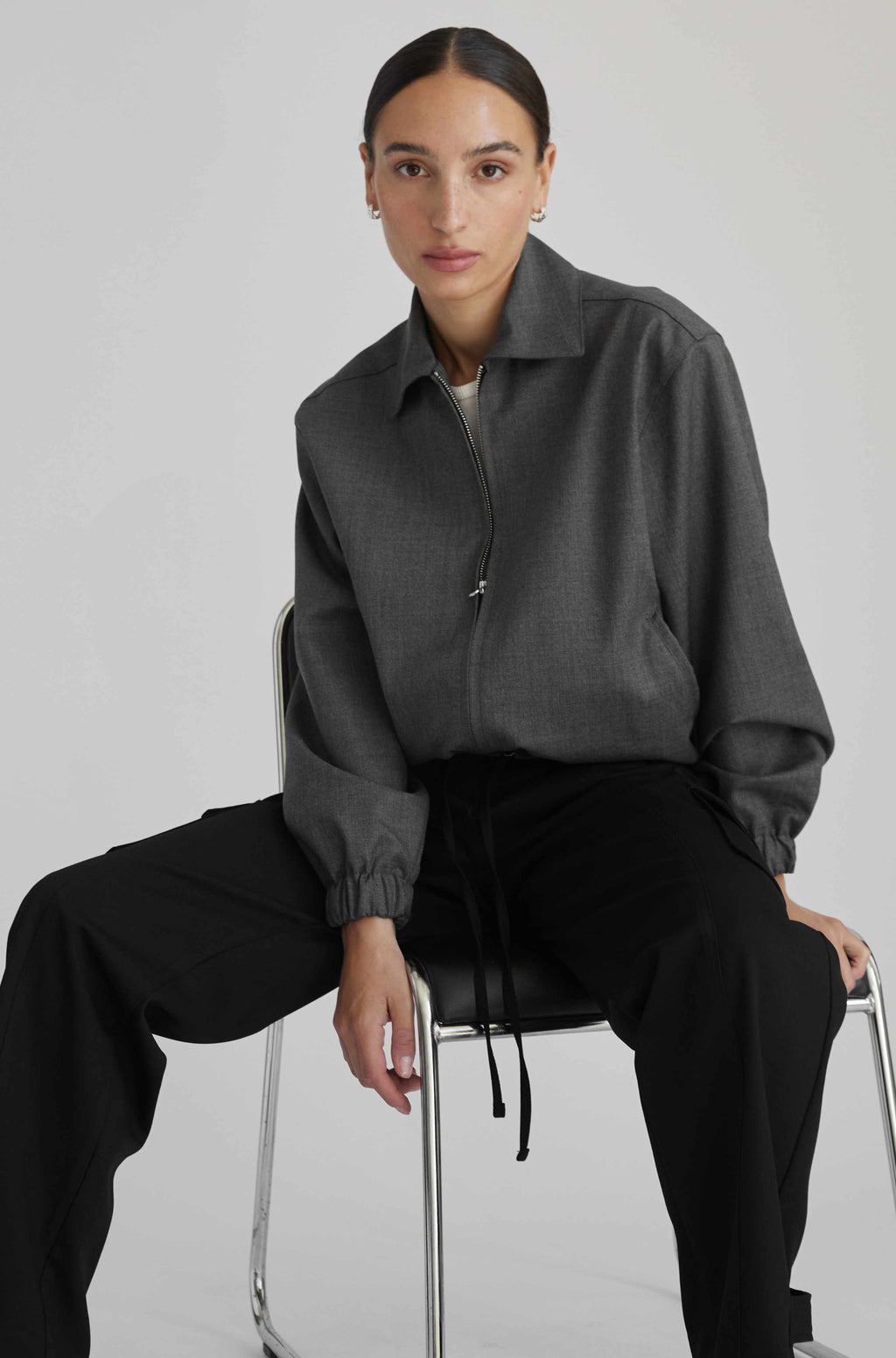 Person wearing a gray jacket and black pants sitting on a stool against a plain background