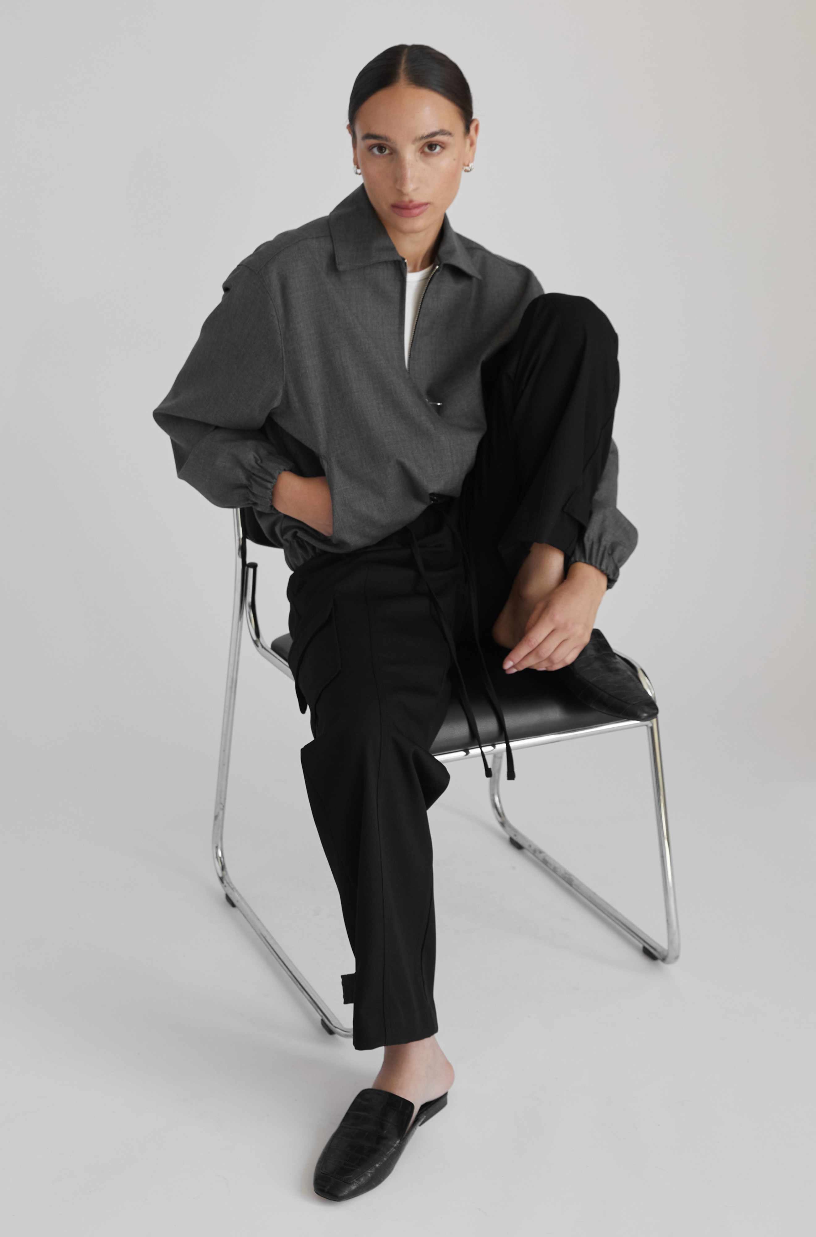 Person wearing a gray shirt and black pants sitting on a chair against a white background