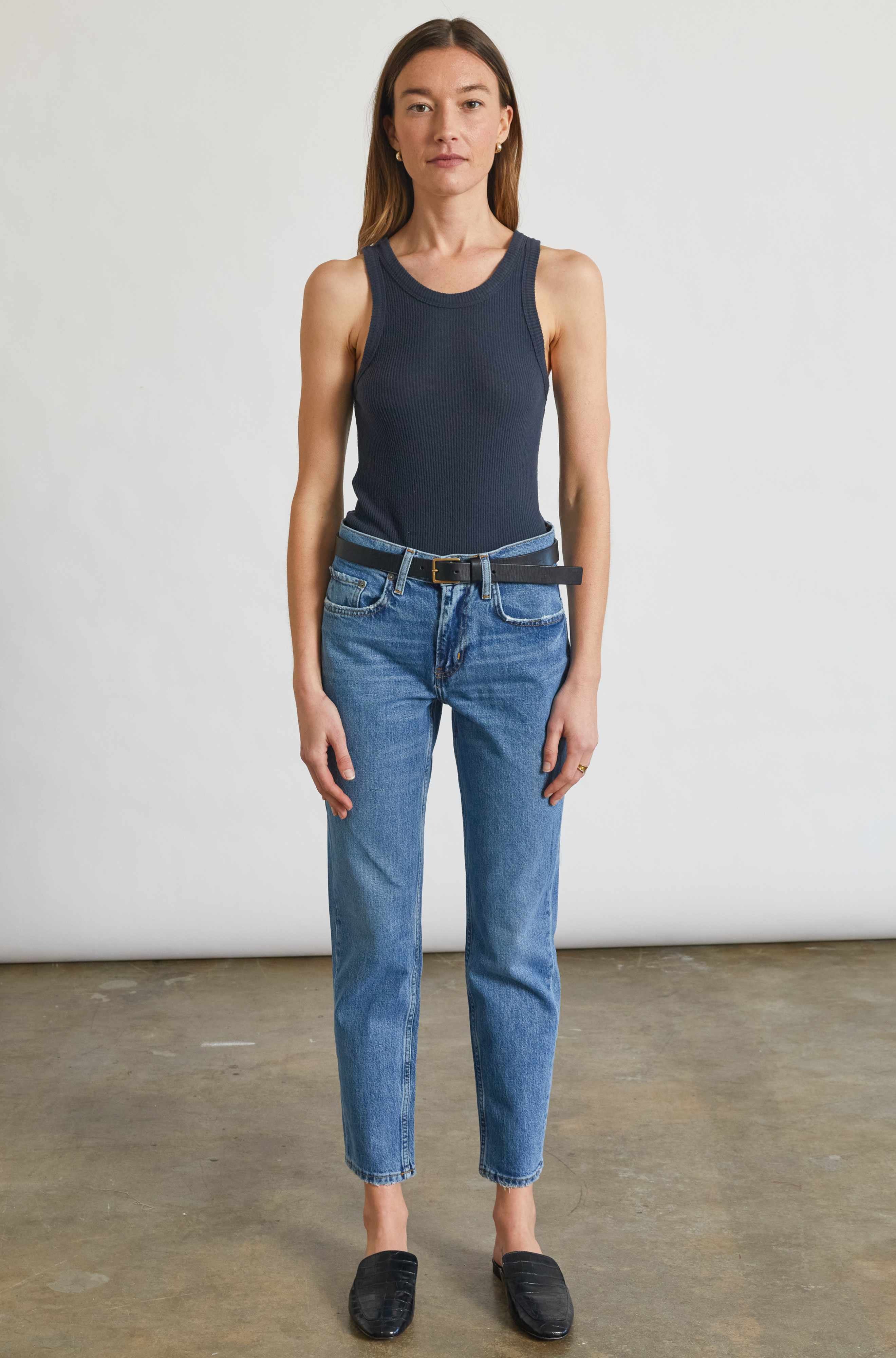 Woman wearing a navy tank top and blue jeans standing against a plain white background