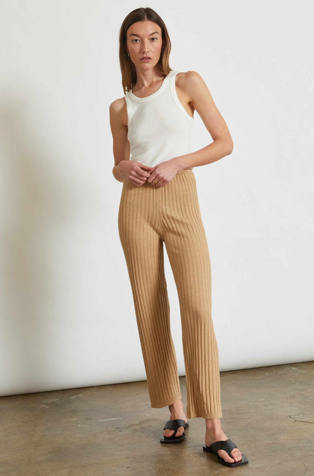 Woman wearing a white sleeveless top and beige striped pants against a plain background