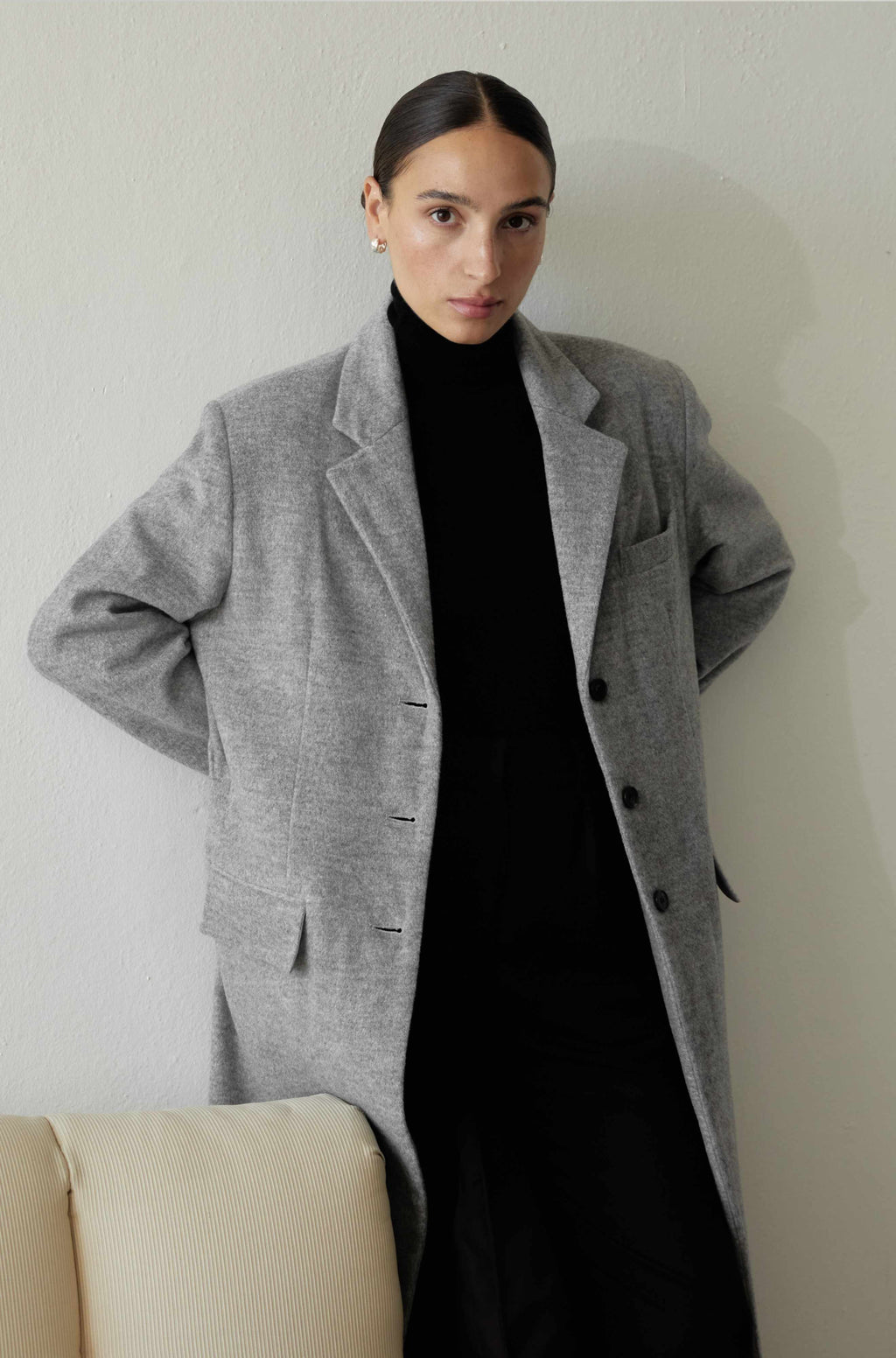 Person wearing a gray coat over a black outfit against a plain background
